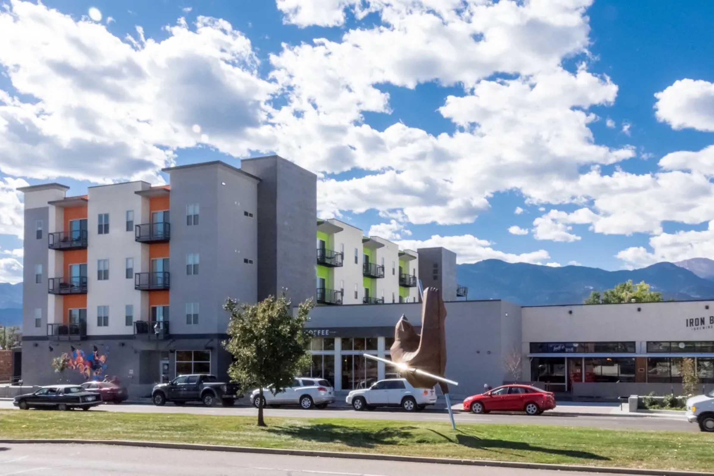 A modern multi-story building with orange and green accents sits alongside a street featuring parked cars. A sculpture of a bird in flight is displayed in the foreground, framed by a backdrop of fluffy clouds and mountains in the distance.
