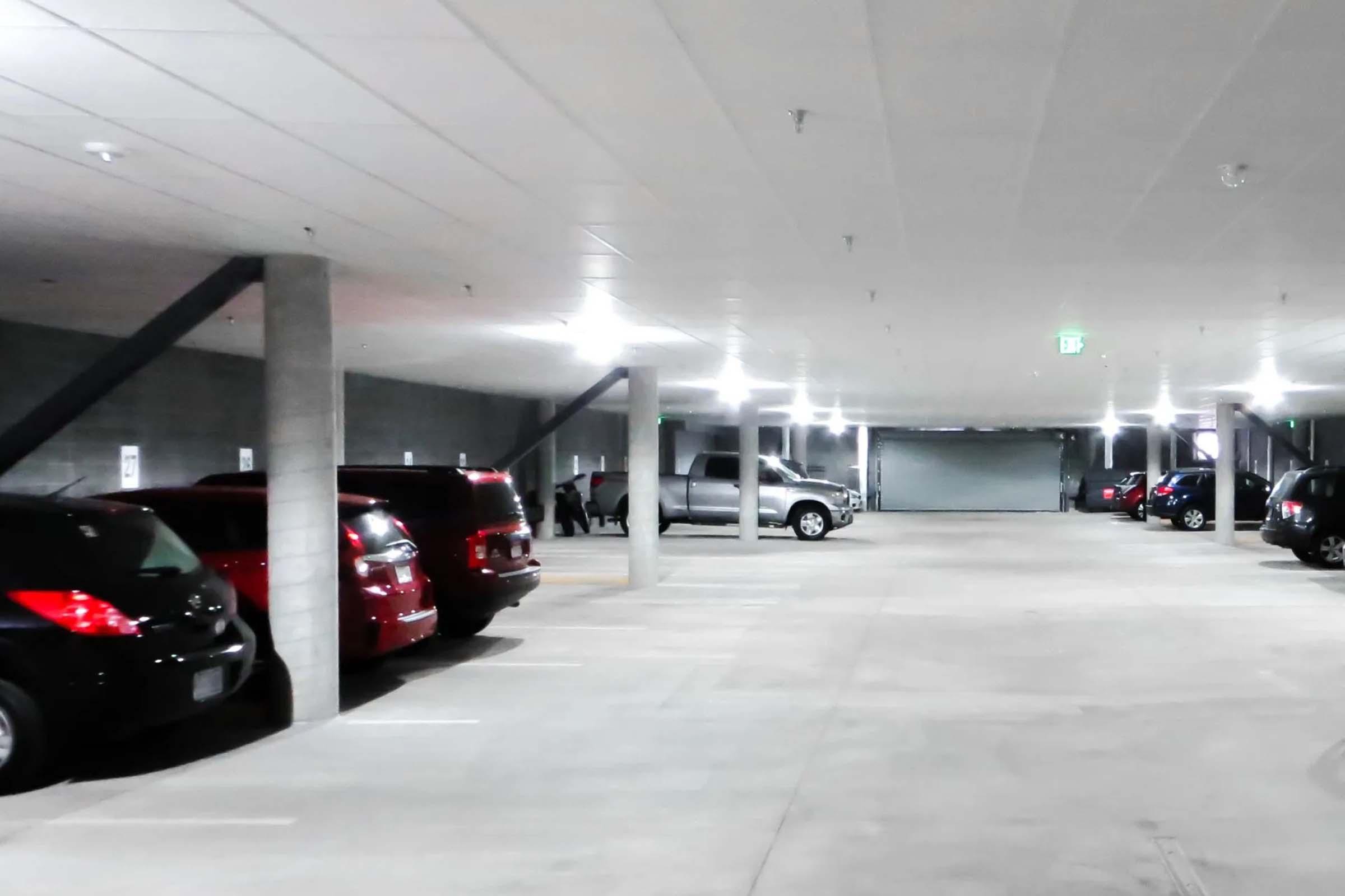 A spacious underground parking garage with rows of parked cars, including red and gray vehicles. The area is well-lit with overhead lights and features concrete pillars. A garage door is visible at the far end, along with a few people in the distance. The floor is smooth and concrete.