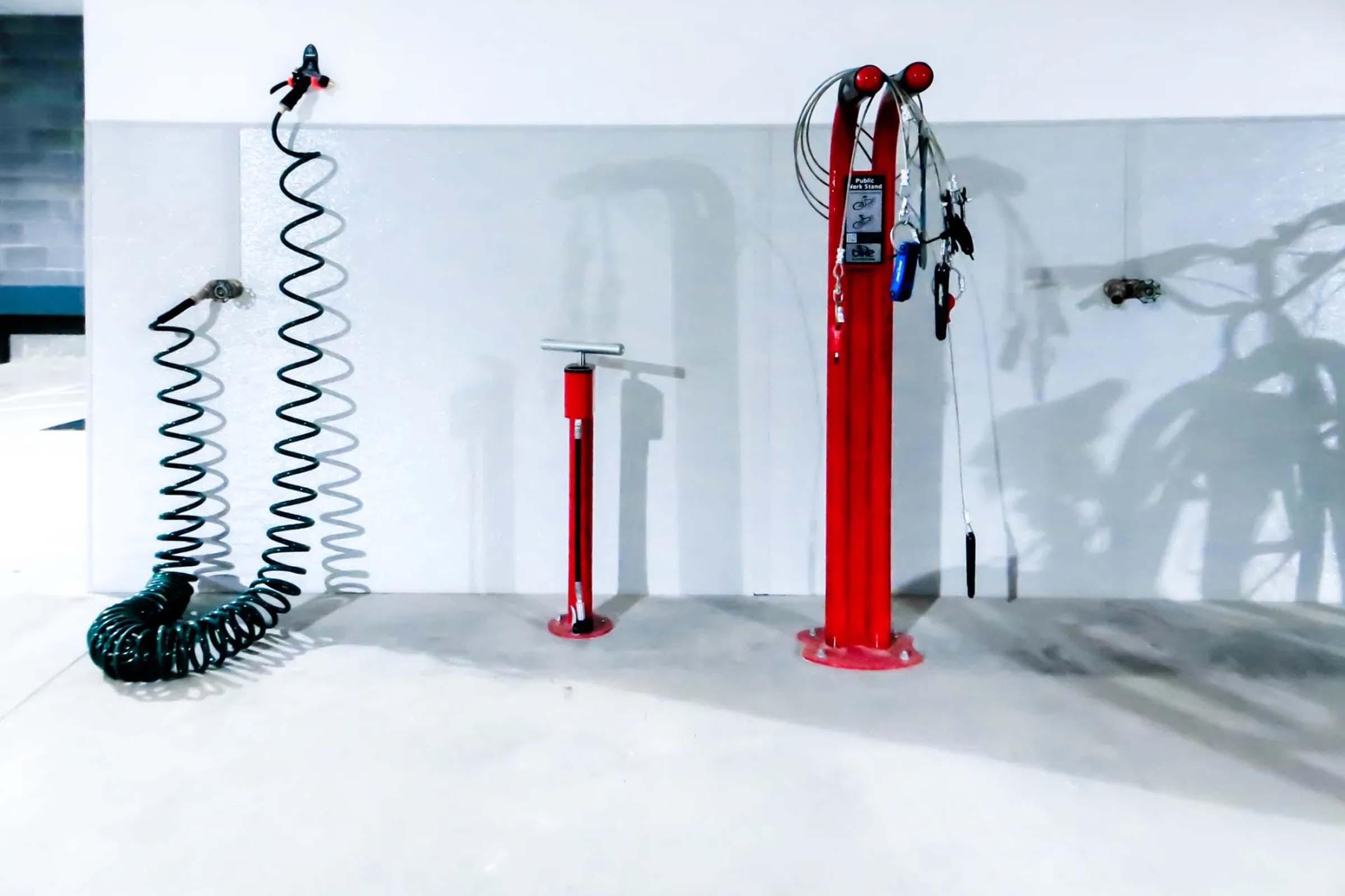 A clean, modern garage or workshop floor featuring three vibrant red air equipment stations. One is a tall air pump with a hose, the second is a shorter inflation device, and the third is a coiled air hose hanging on the wall, with tools neatly arranged nearby. The background is minimalistic with a gray wall.