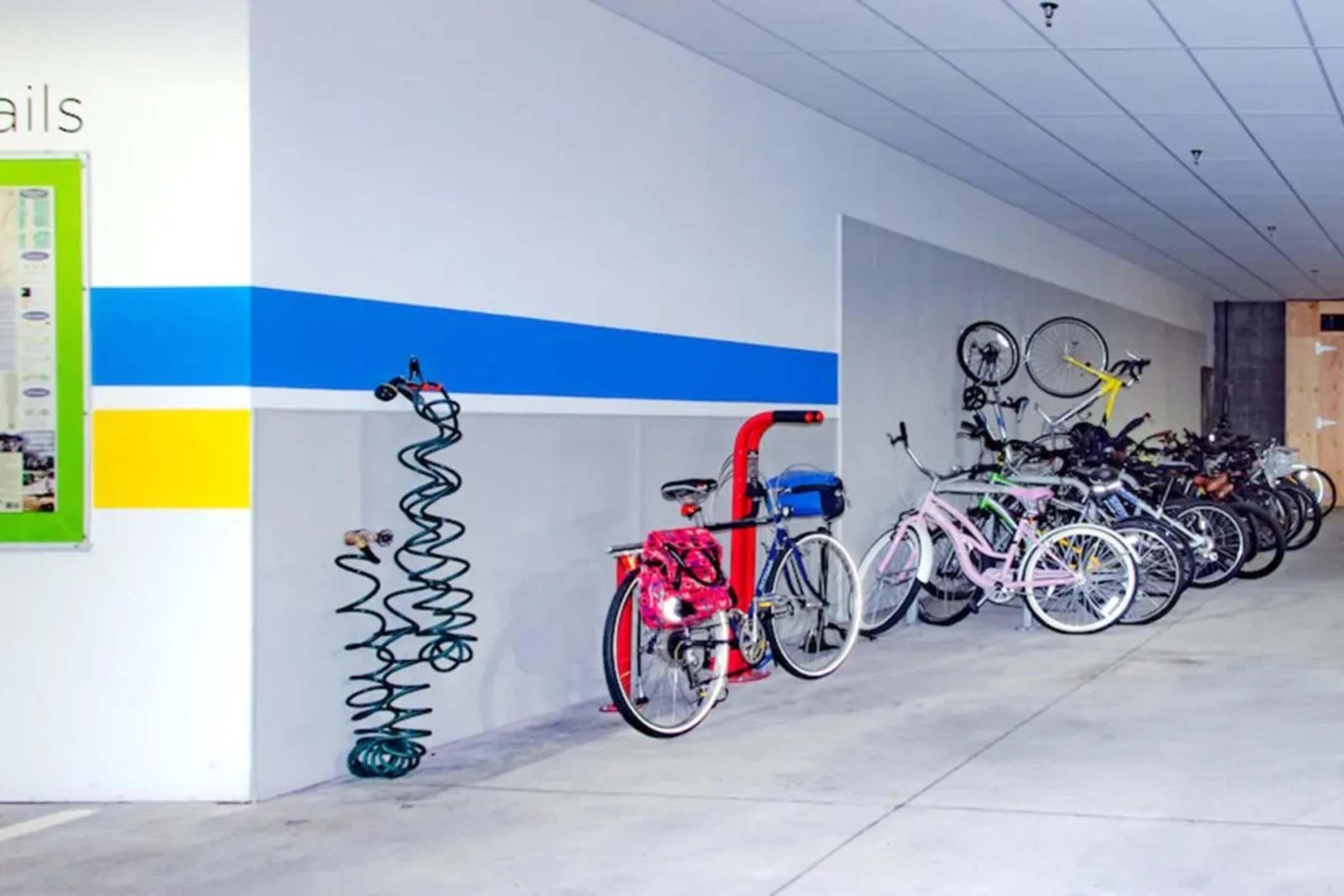A bicycle storage area featuring various bikes parked along a wall. The space includes a bike repair station with tools and colorful bike racks. The wall is painted with blue and yellow stripes, and there are a few decorative elements alongside the bicycles.