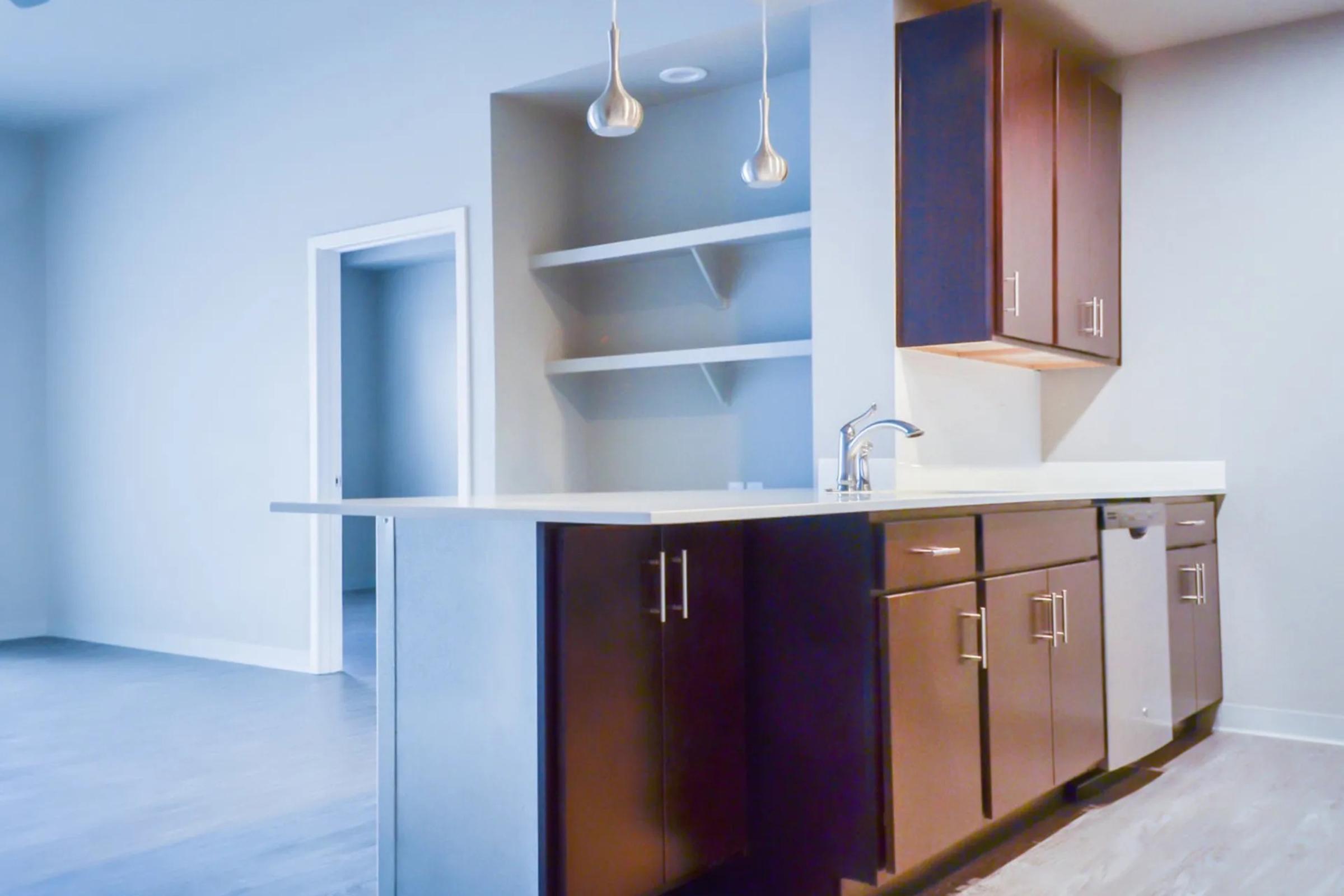 Modern kitchen with dark wood cabinets and a sleek countertop. Two pendant lights hang above the breakfast bar. The room features an open layout with light-colored walls and a door leading to another space, enhancing a spacious feel.