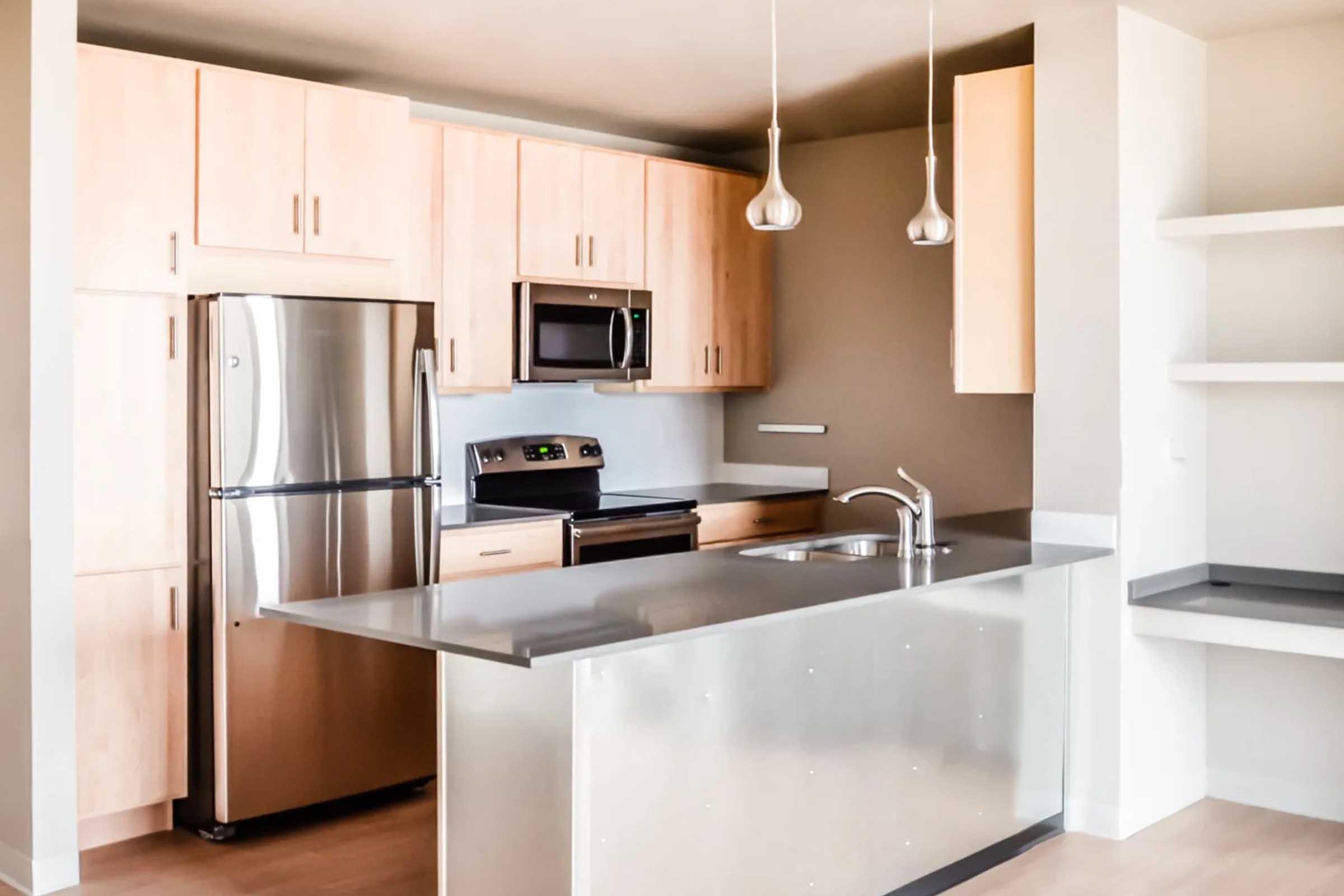 Modern kitchen featuring light wood cabinetry, stainless steel appliances including a refrigerator and microwave, a sleek silver countertop, and pendant lighting. The design emphasizes a clean, minimalist aesthetic with an open layout and neutral color scheme.