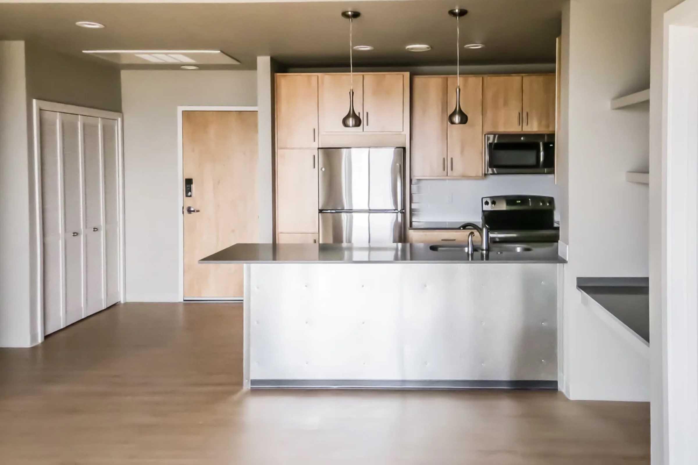 Modern kitchen area featuring stainless steel appliances, including a refrigerator and microwave, alongside wooden cabinets and a sleek countertop. The open layout leads to a living space with light-colored walls and hardwood flooring, providing a bright and inviting atmosphere.
