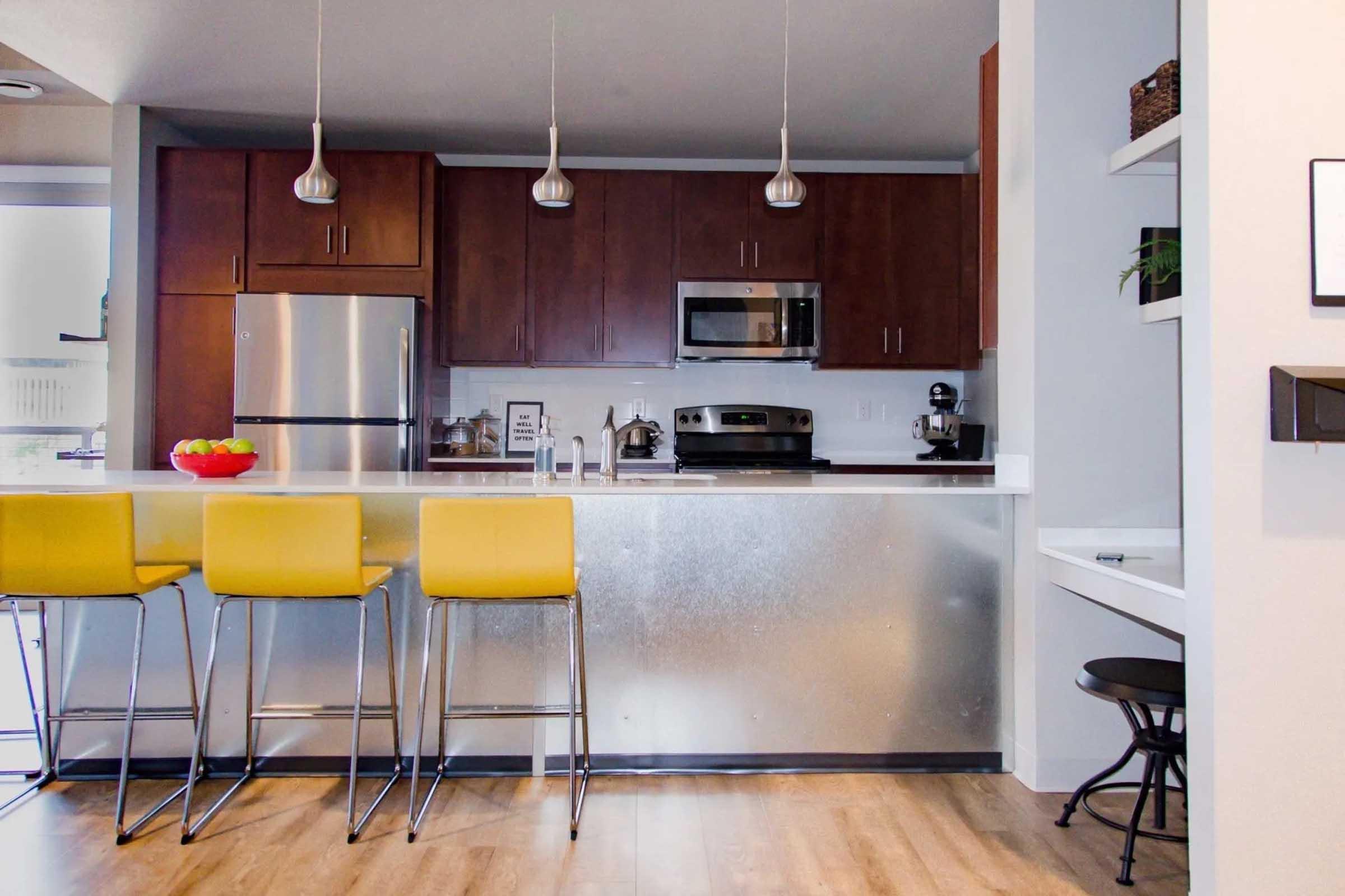 Modern kitchen featuring stainless steel appliances, dark wood cabinetry, and a sleek island with three yellow bar stools. A fruit bowl sits on the counter, and a small nook is visible to the side, creating a stylish and functional cooking space. Bright and inviting atmosphere with natural light.
