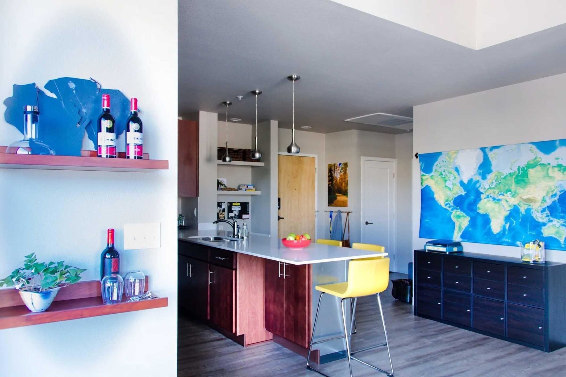 A modern kitchen featuring a central island with yellow bar stools, wine bottles on a shelf, and various kitchen items. A large world map is mounted on the wall, adding a vibrant touch to the space. The overall design is sleek, with wooden cabinets and subtle decor, creating a welcoming atmosphere.