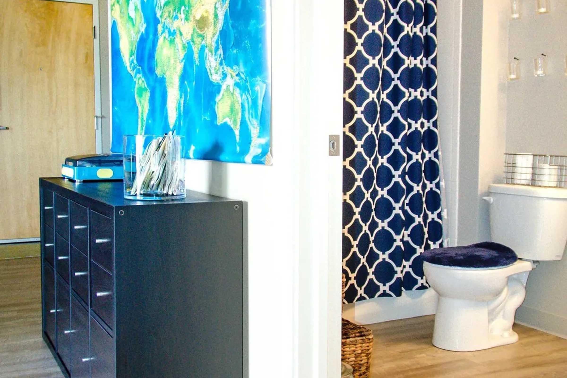 A modern interior space featuring a large world map on the wall. A dark storage unit sits beside a bathroom area, which includes a white toilet and a decorative shower curtain with a navy geometric pattern. The flooring is light wood, and the overall design is clean and contemporary.