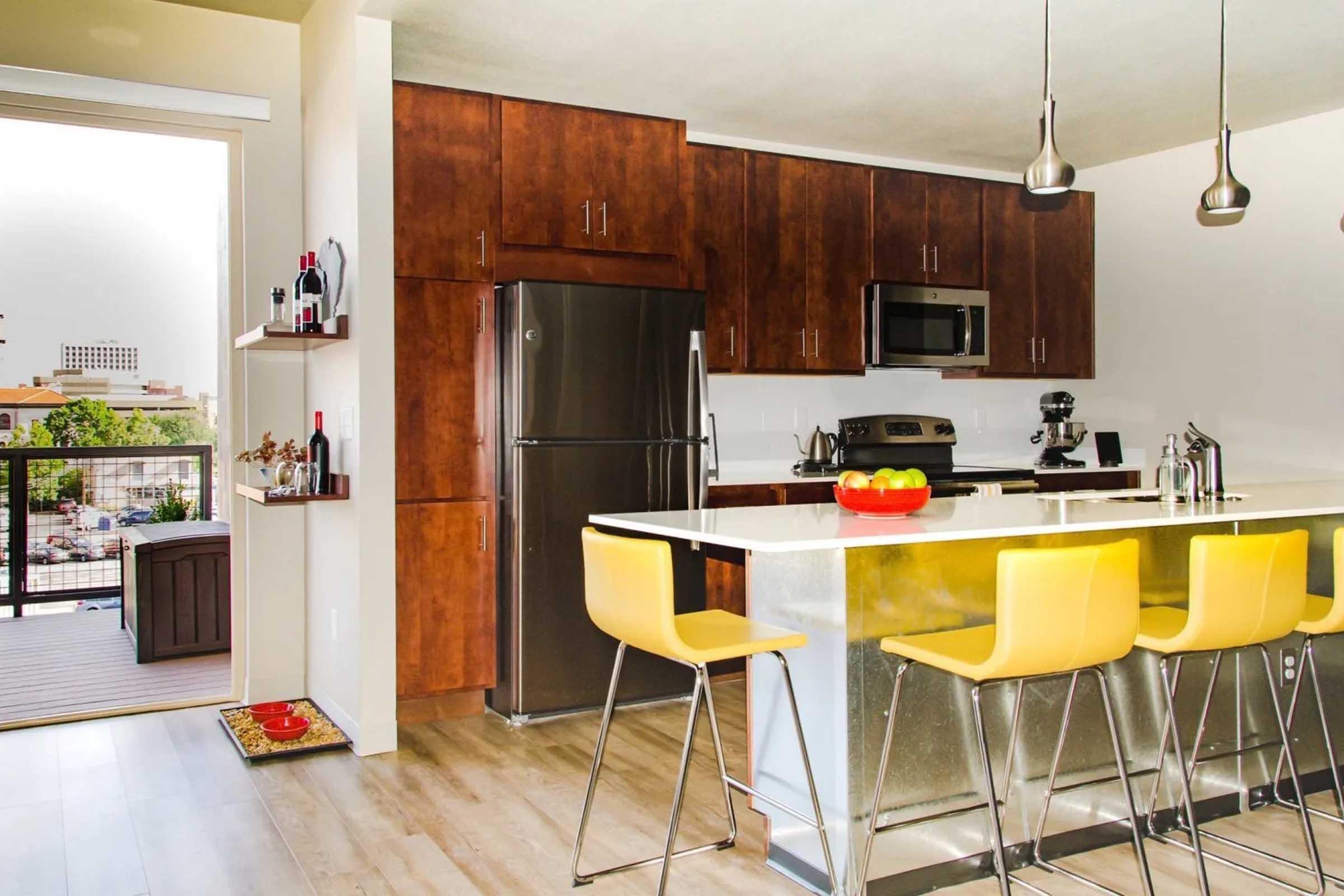 Modern kitchen featuring dark wood cabinets, stainless steel appliances, and a large island with yellow bar stools. The countertop is bright and glossy, and there are bottles on a nearby shelf. A sliding door opens to a balcony, allowing natural light to enter the space.