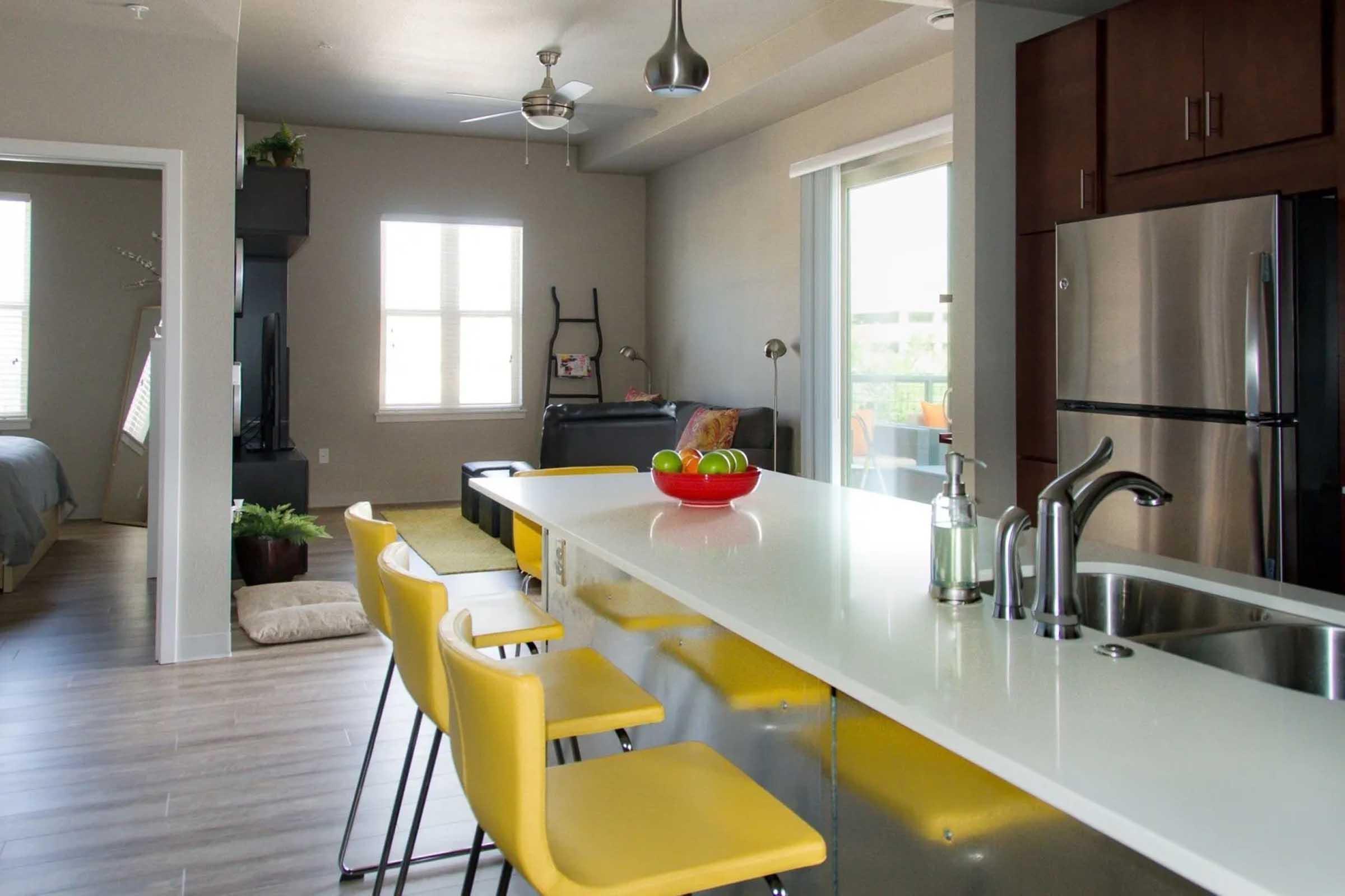 A modern kitchen featuring a sleek white countertop with yellow bar stools. In the background, a cozy living area is visible, along with a bedroom space. Natural light floods the room through large windows, highlighting the contemporary furniture and warm color palette.