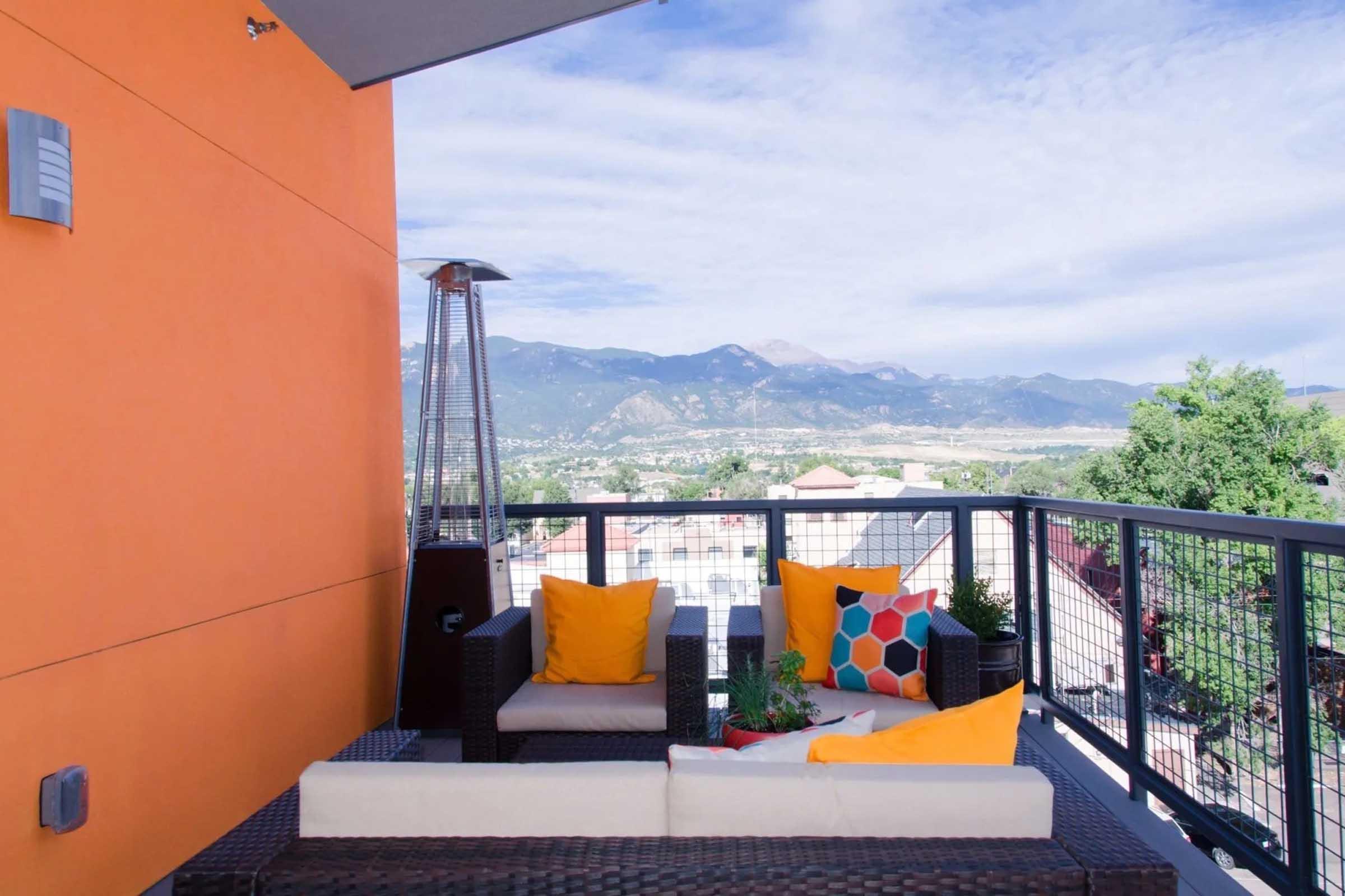 A modern balcony with wicker furniture featuring bright orange cushions and decorative pillows. In the background, mountains and a clear blue sky create a scenic view, while a heater adds a cozy touch. The wall is painted in an eye-catching orange hue, enhancing the vibrant outdoor atmosphere.