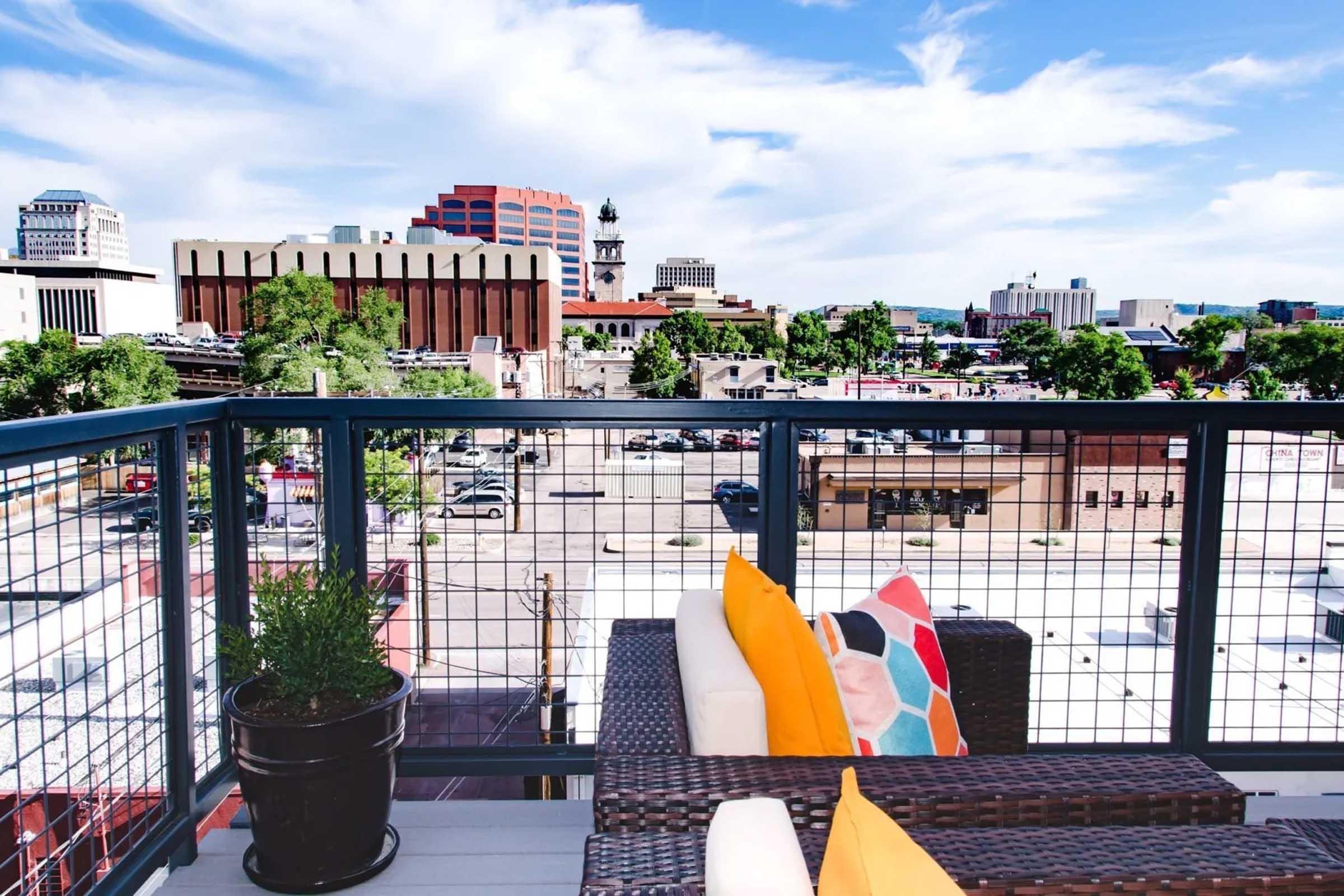 A scenic view from a balcony featuring a modern railing and a wicker seating area with colorful pillows. In the background, city buildings and a blue sky with scattered clouds are visible, showcasing an urban landscape. A potted plant adds a touch of greenery.