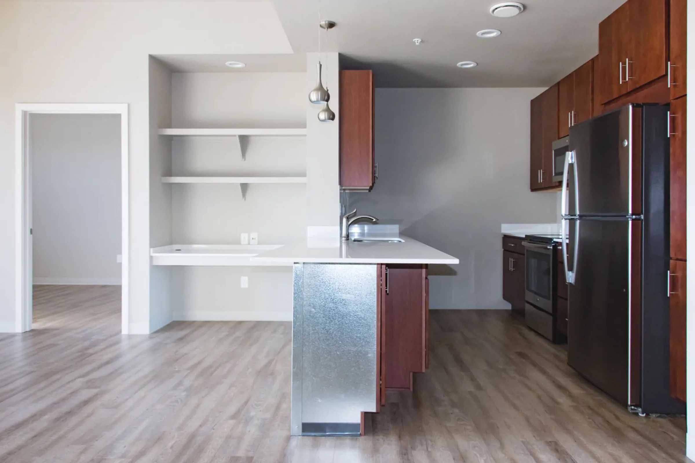 A modern kitchen featuring dark wood cabinets, stainless steel appliances, and a large countertop island. The space includes a silver refrigerator, a white sink, and a contrasting light gray wall. To the left, there is an open area leading to another room with shelves. The flooring is light wood.