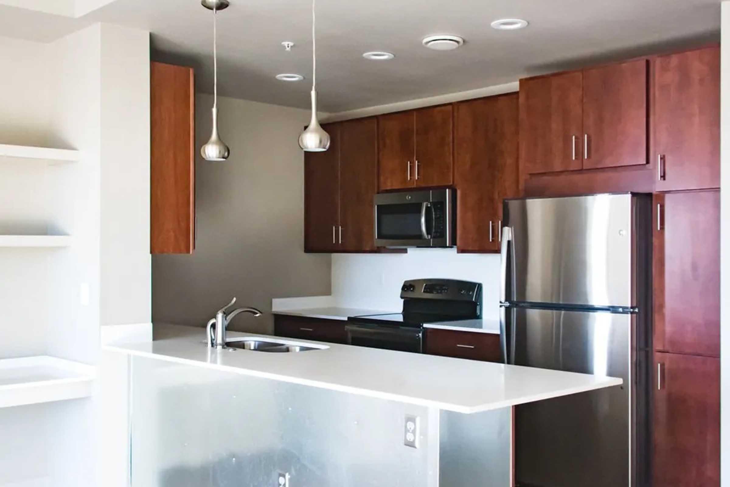 Modern kitchen featuring wooden cabinets, stainless steel appliances including a microwave, an oven, and a fridge. The countertop is white, with a sink and space for seating at a bar-style ledge. The walls are neutral-colored, creating a clean and contemporary look.
