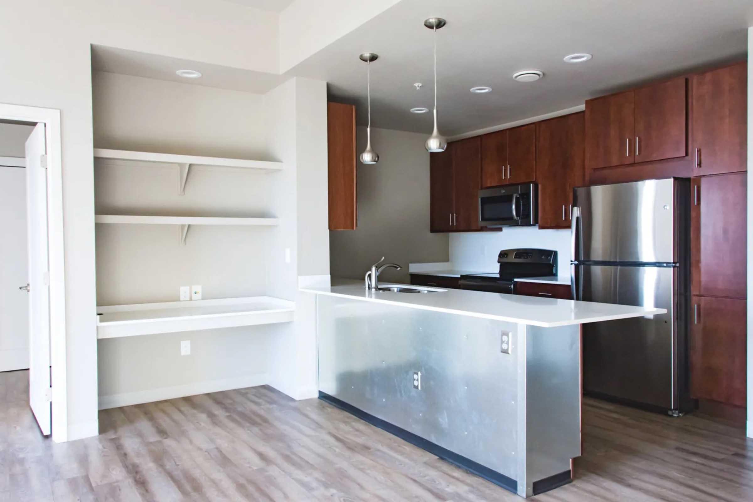 Modern kitchen with wooden cabinets, stainless steel appliances, and a central island. Features include a built-in shelf, sleek countertop, and pendant lighting. The flooring is a light wood, enhancing the bright and airy feel of the space. Perfect for a contemporary living environment.
