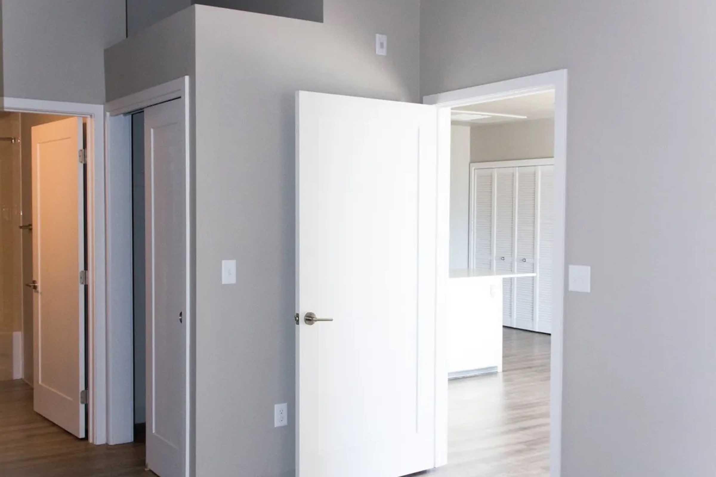 A modern interior featuring a light gray wall with a doorway leading to a small room. There are two closed white doors, one leading to a closet or bathroom, and another to another room. The flooring is a light wood, and natural light is coming from the left, illuminating the space.