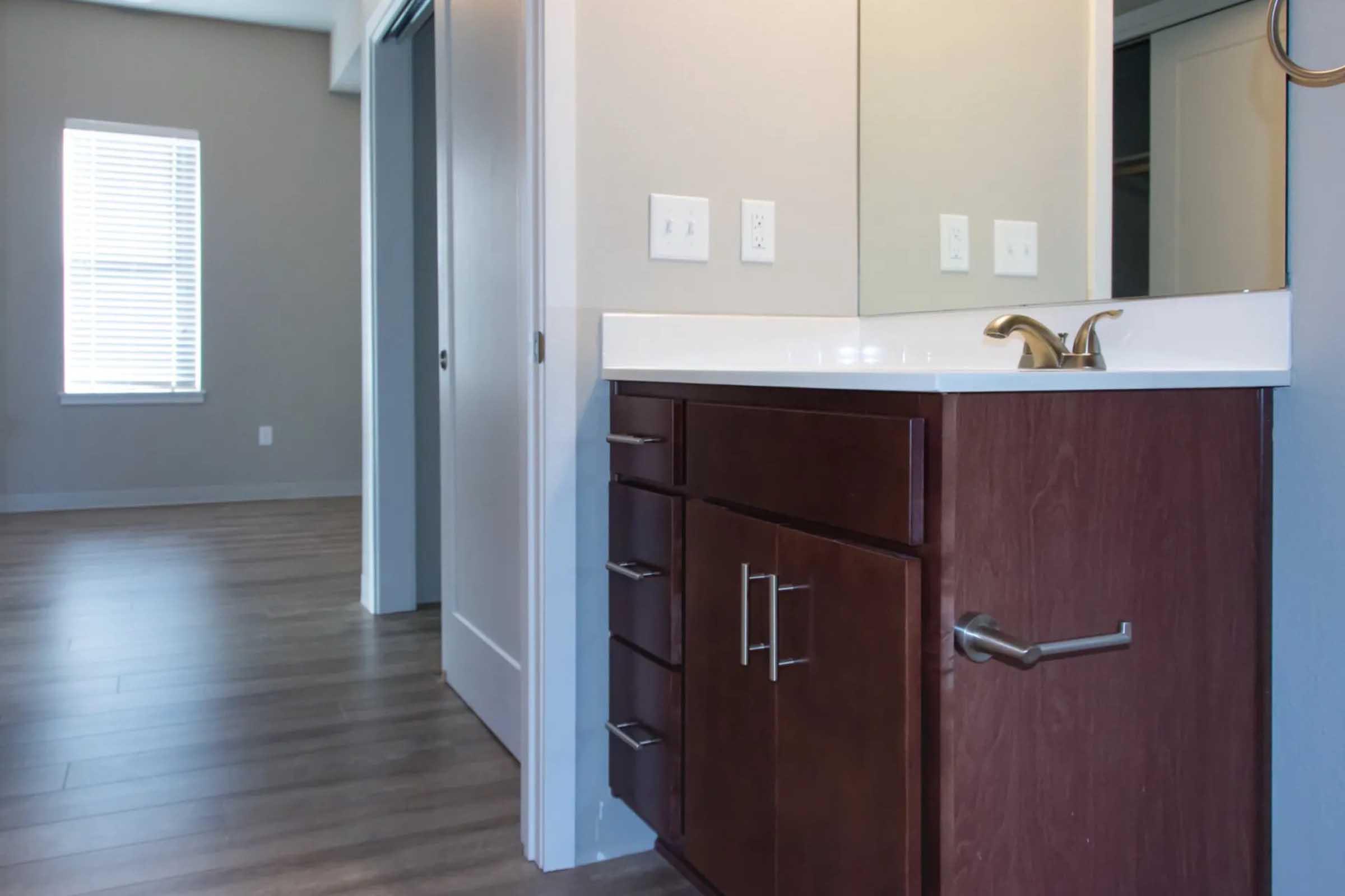 A modern bathroom featuring a dark wood vanity with multiple drawers, a white countertop, and a single sink. There is a large mirror above the sink, and the room is well-lit by natural light from a nearby window. The flooring is a light-colored wood, creating a clean and contemporary look.