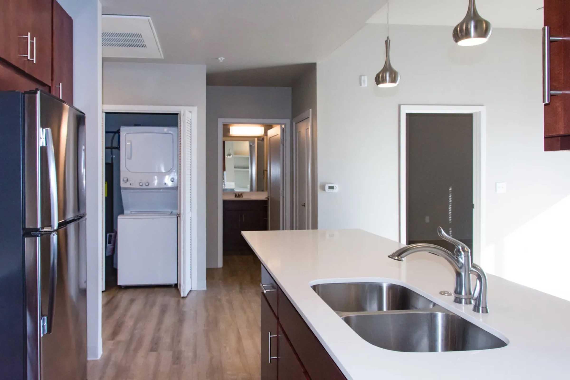 A modern kitchen featuring a stainless steel refrigerator and a double sink with a sleek faucet. The open layout includes wooden cabinetry and a view of a laundry area and bathroom through adjacent doorways. Natural light floods the space, enhancing the contemporary design.