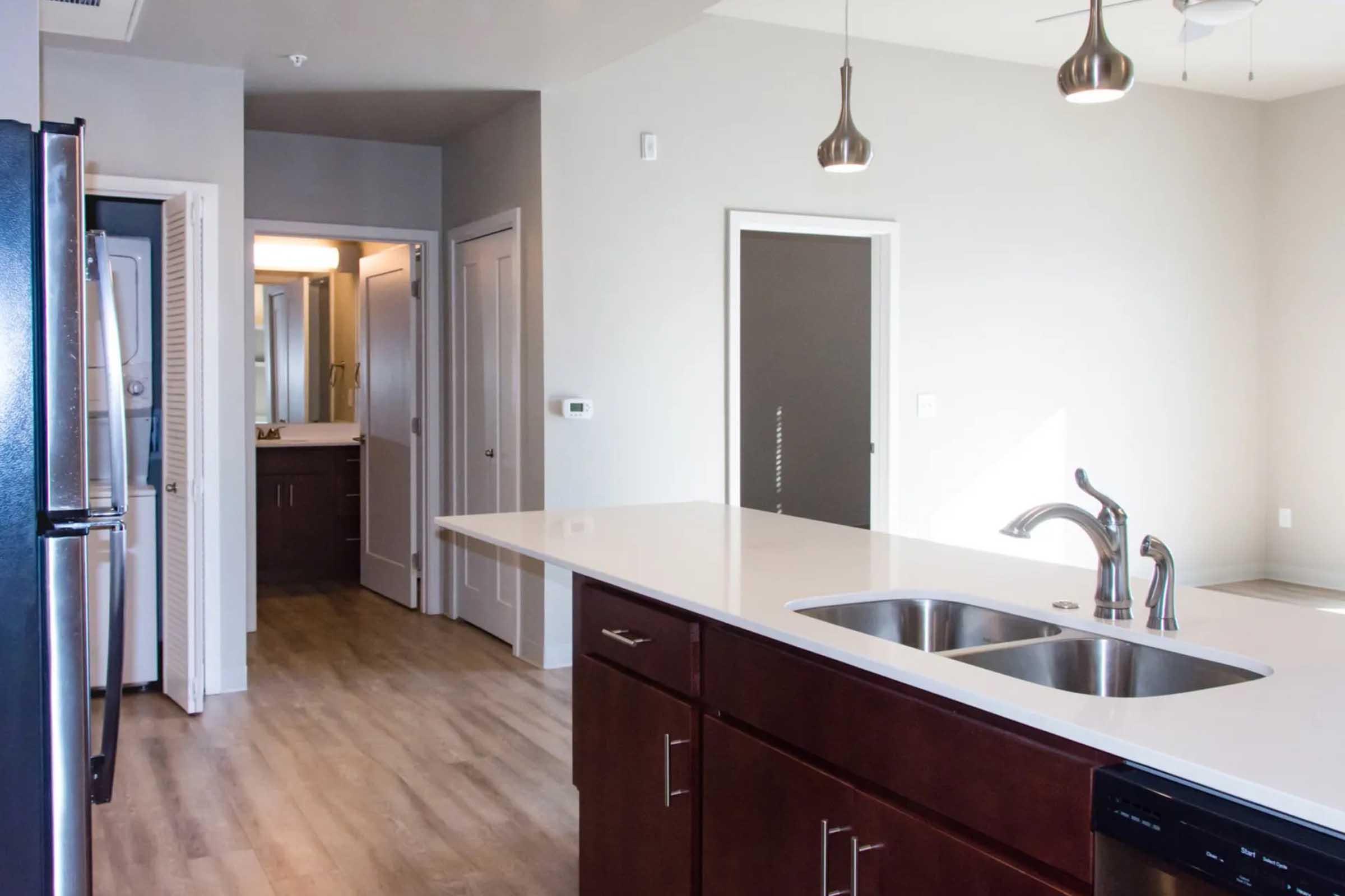 Modern kitchen with dark wood cabinetry and a white countertop featuring a double sink. To the left, there is a stainless steel refrigerator. In the background, an open doorway leads to a hallway with additional doors, and there are pendant lights hanging from the ceiling. The flooring is light-colored wood.