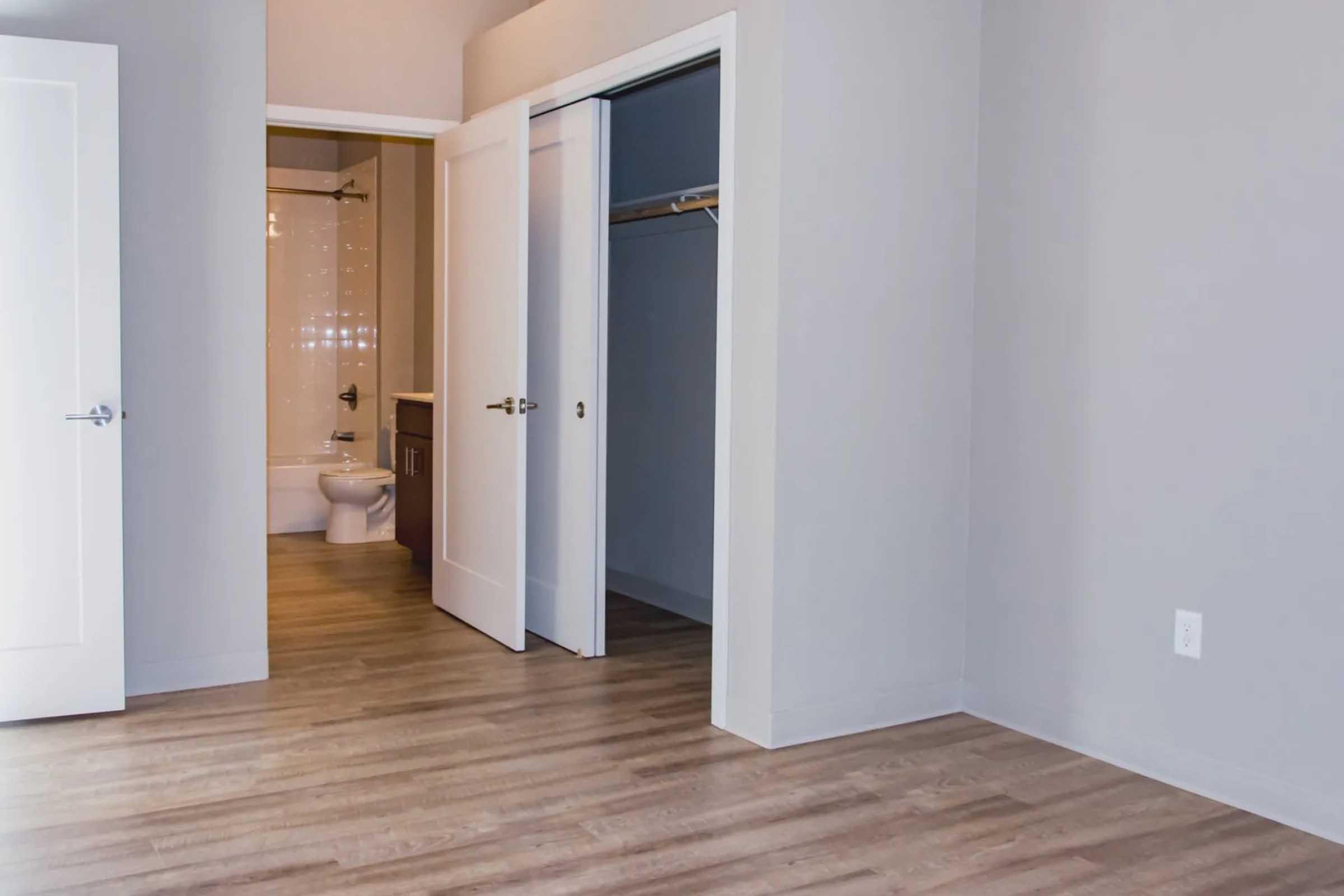 A view of a light-filled room with grey walls and wooden flooring. Two white doors are partially open, revealing a closet and a bathroom area in the background. The bathroom features white tiles and a toilet, while the room appears empty and unadorned, giving it a clean, modern aesthetic.