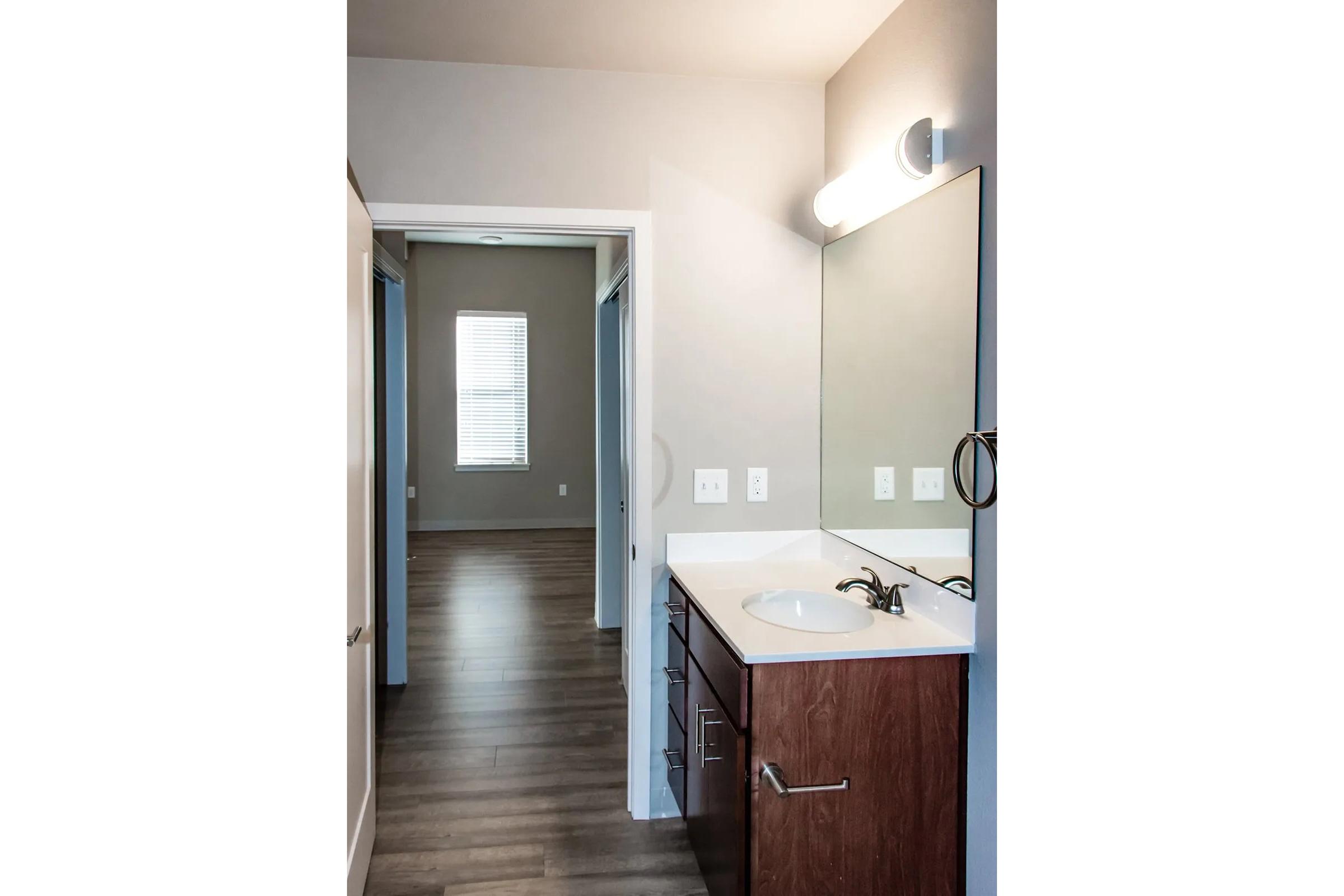 A modern bathroom interior featuring a sink with a dark wood vanity, a large mirror above it, and a wall-mounted light fixture. The room adjoins a hallway leading to another space with windows, showcasing light wood flooring and neutral wall colors.