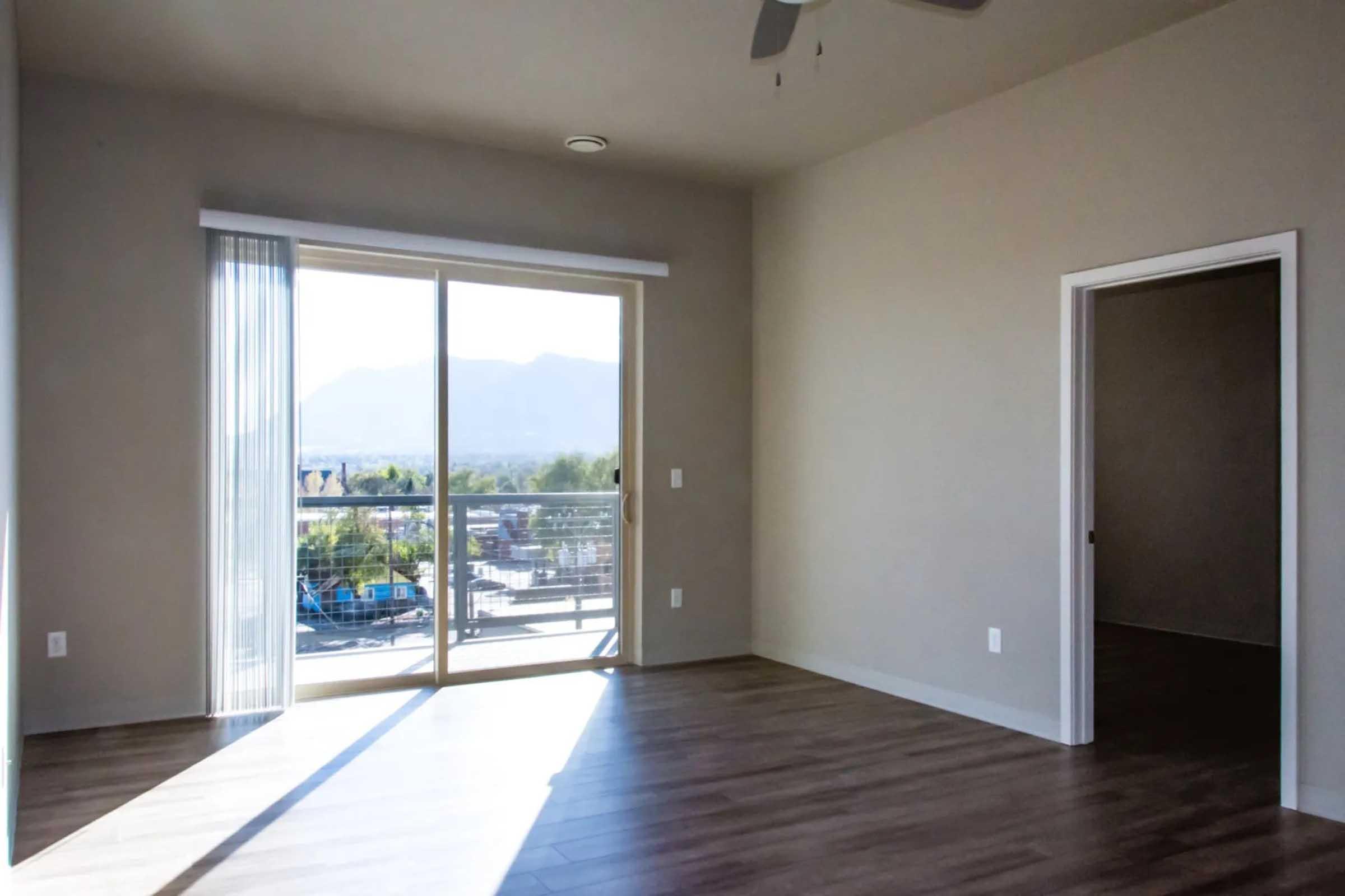 Bright and airy room with large sliding glass doors leading to a balcony, offering a view of mountains. The floor is finished with light wood, and the walls are painted in neutral tones. A doorway to another room is visible on the right. Sunlight fills the space, creating a welcoming atmosphere.