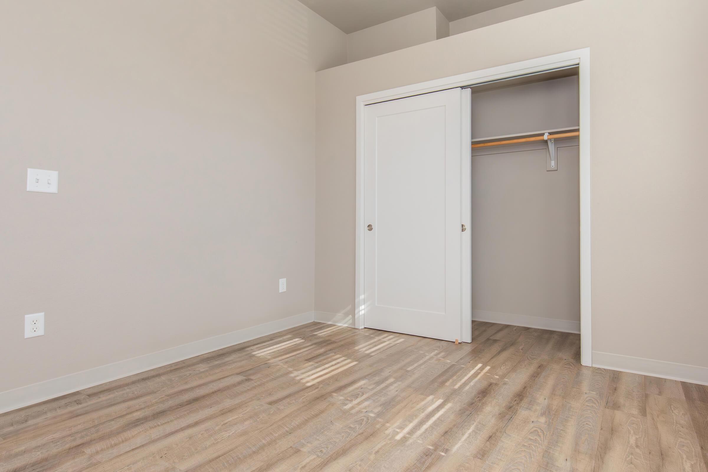 A bright, unfurnished room with light-colored walls and wooden flooring. A white closet with a sliding door is visible on the right, featuring a wooden rod for hanging clothes. Natural light streams in from a window, creating a warm atmosphere.