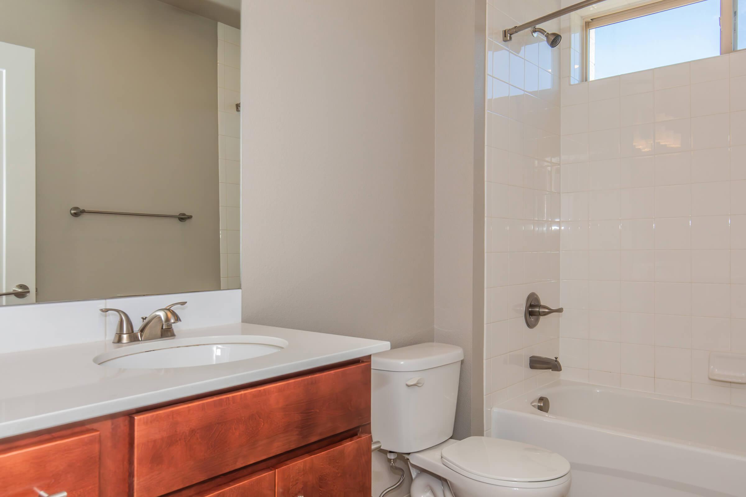 A modern bathroom featuring a white tile shower with a bathtub, a wooden vanity with a sink, and a toilet. Natural light enters through a window, illuminating the space, which has neutral-colored walls and simple fixtures. A towel rack is mounted on the wall beside the mirror.