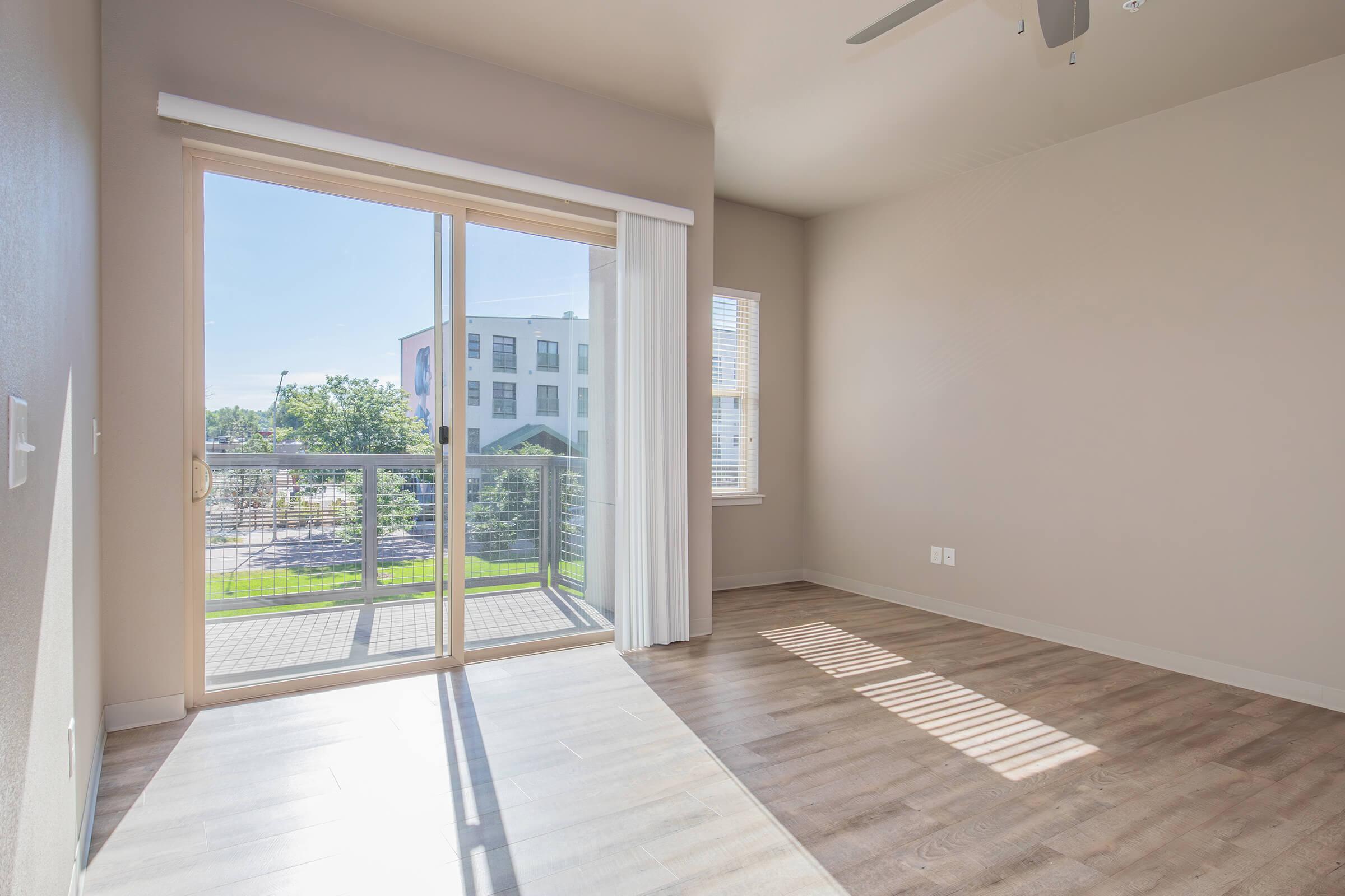 A bright, empty room with large sliding glass doors leading to a balcony. Natural light floods the space, highlighting light-colored walls and wooden flooring. In the background, a glimpse of greenery and buildings can be seen through the balcony.
