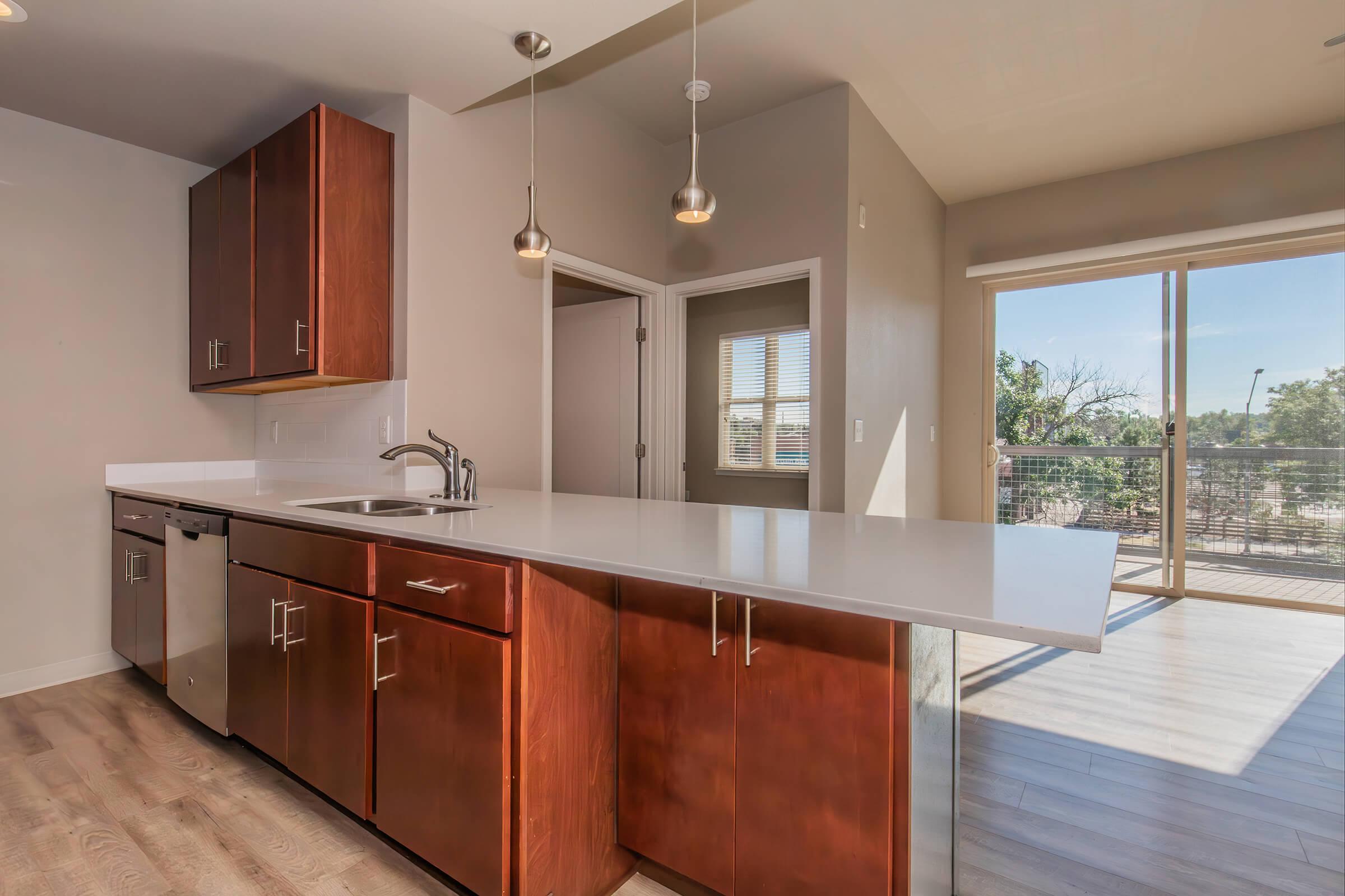 Modern kitchen featuring a sleek design with dark wood cabinets, a stainless steel dishwasher, and a minimalist countertop. Pendant lights hang over the kitchen island, and large windows provide natural light, showcasing a view of the outdoors. The space has an open layout, connecting to a living area.