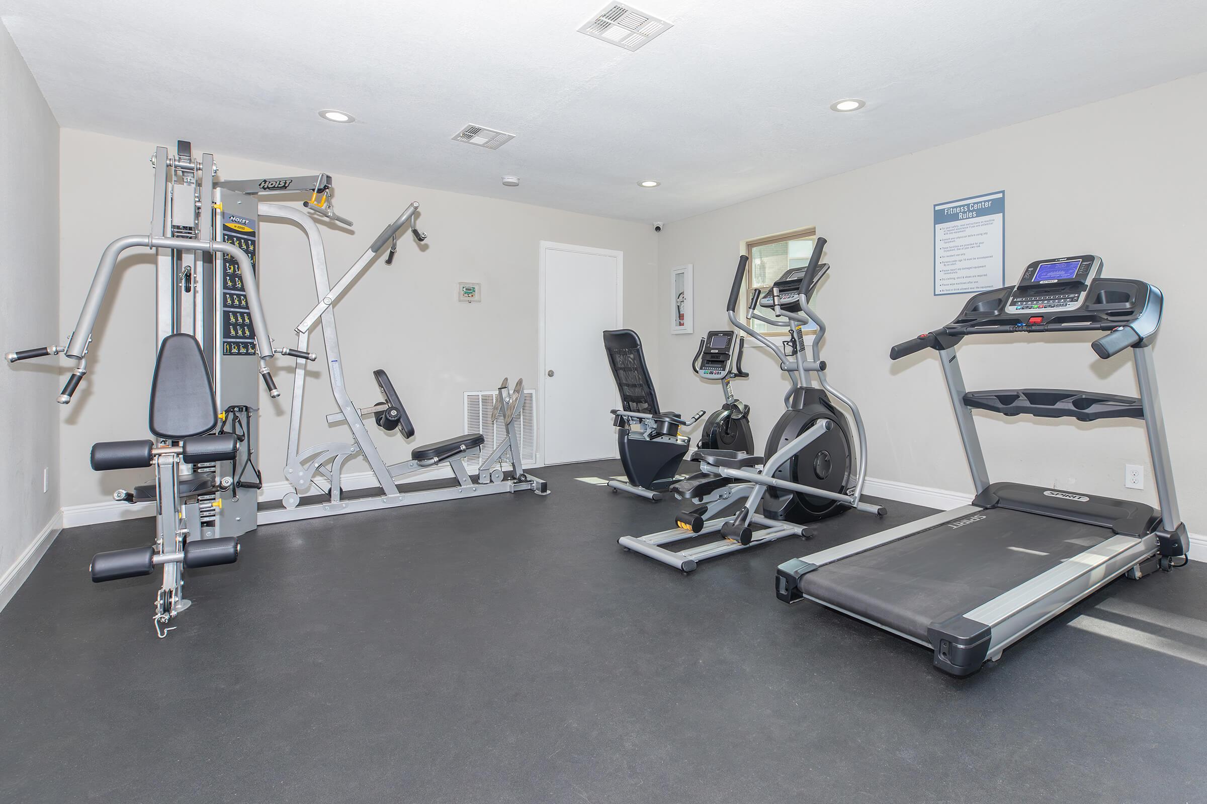 A modern home gym featuring various workout equipment, including a multi-station strength machine, an elliptical trainer, a recumbent exercise bike, and a treadmill, set against a neutral wall with minimal decor and good lighting.