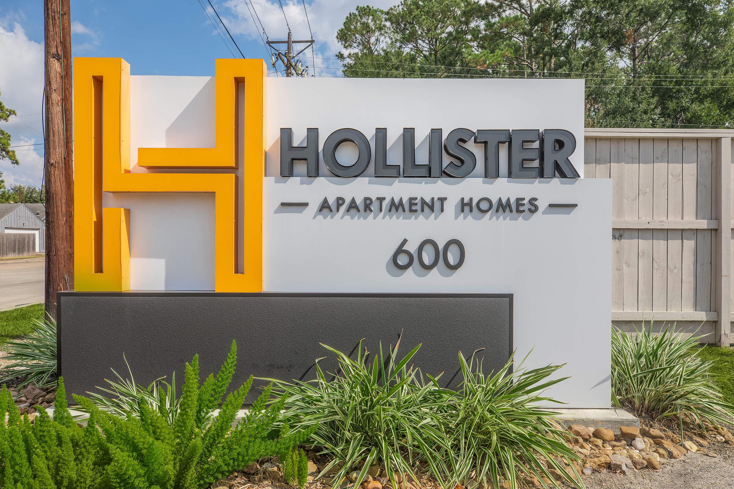 Sign for Hollister Apartment Homes featuring a large orange "H" and the text "600" below. Surrounded by greenery, the sign is set against a clear blue sky with a wooden fence in the background.
