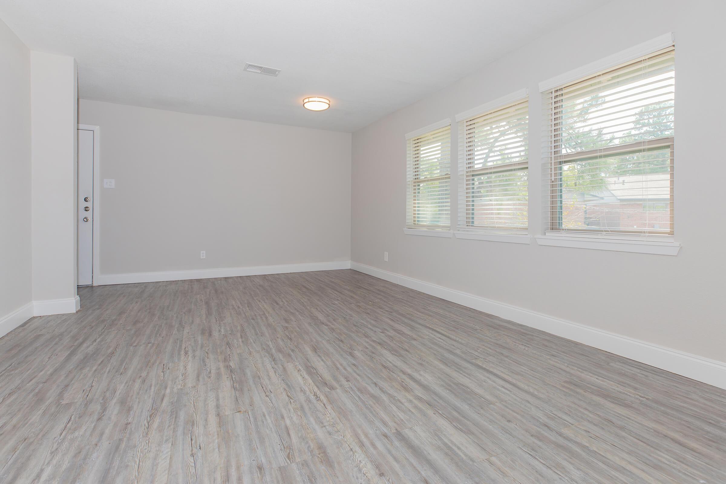 A spacious, empty living room featuring light gray walls and large windows with blinds. The floor is covered in wood-like laminate, providing a modern look. A ceiling light fixture is visible, and there's a door leading to another room. The overall ambiance is bright and inviting.