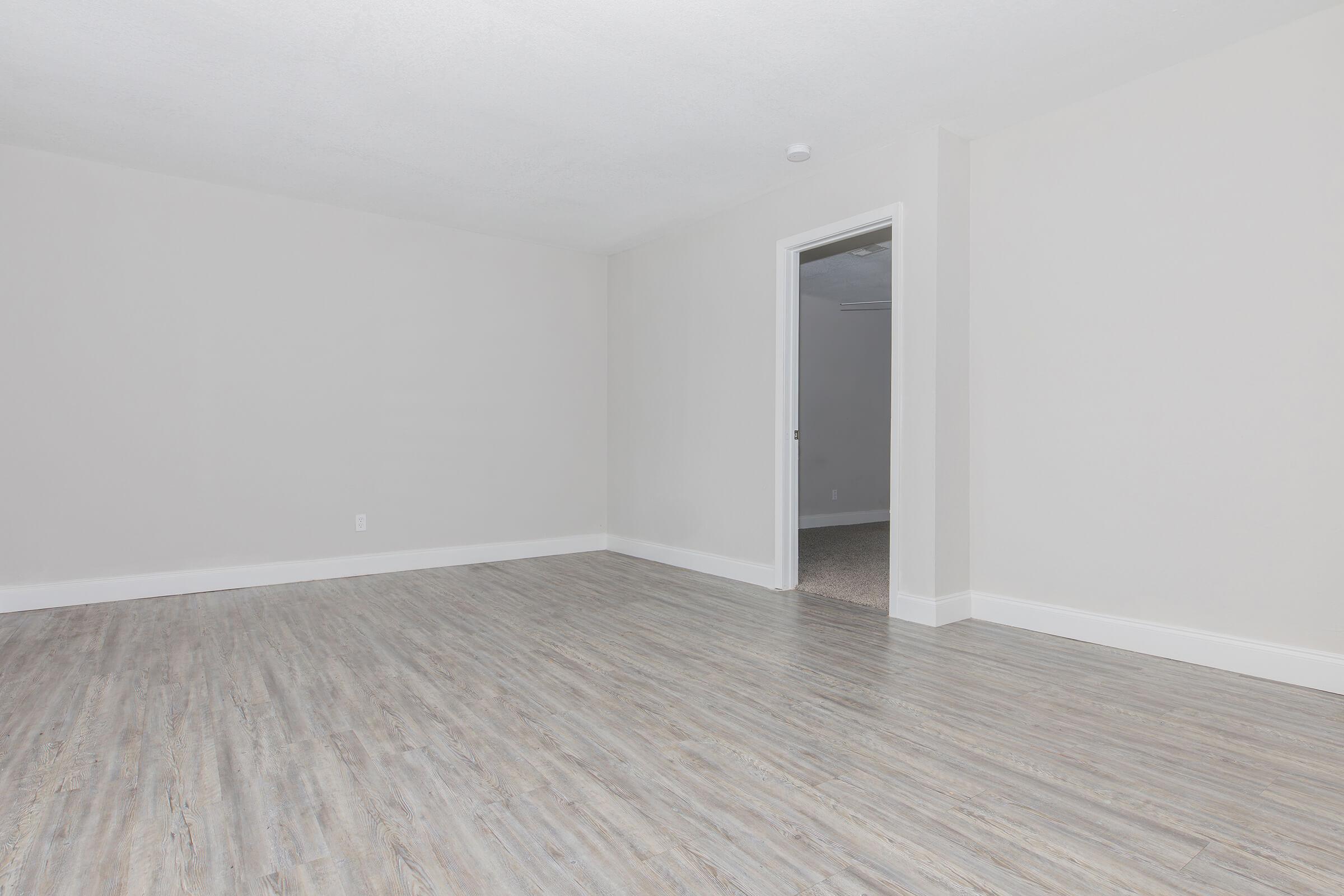 A spacious, empty room with light gray walls and a light wood floor. There is a door leading to another room visible in the background, creating an inviting and clean space with ample natural light. The overall atmosphere is minimalistic and airy.
