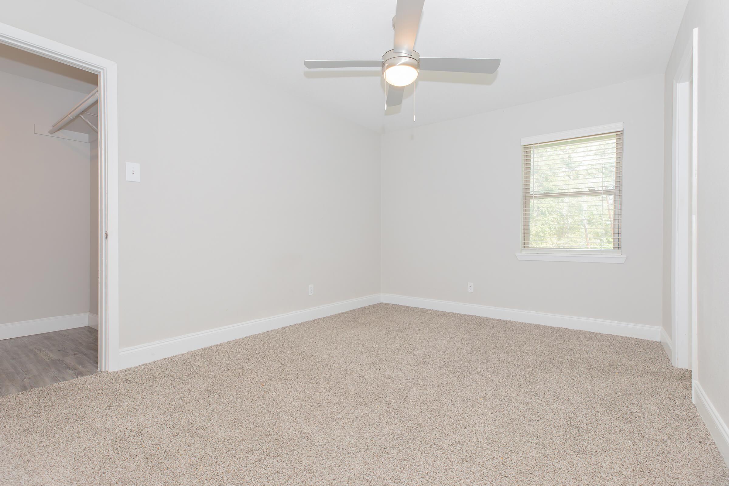 A sparsely furnished room with light gray walls and beige carpeting. It features a ceiling fan and a window with blinds allowing natural light. In the background, there is an open closet space. The overall appearance is clean and well-lit, ideal for a living space or bedroom.