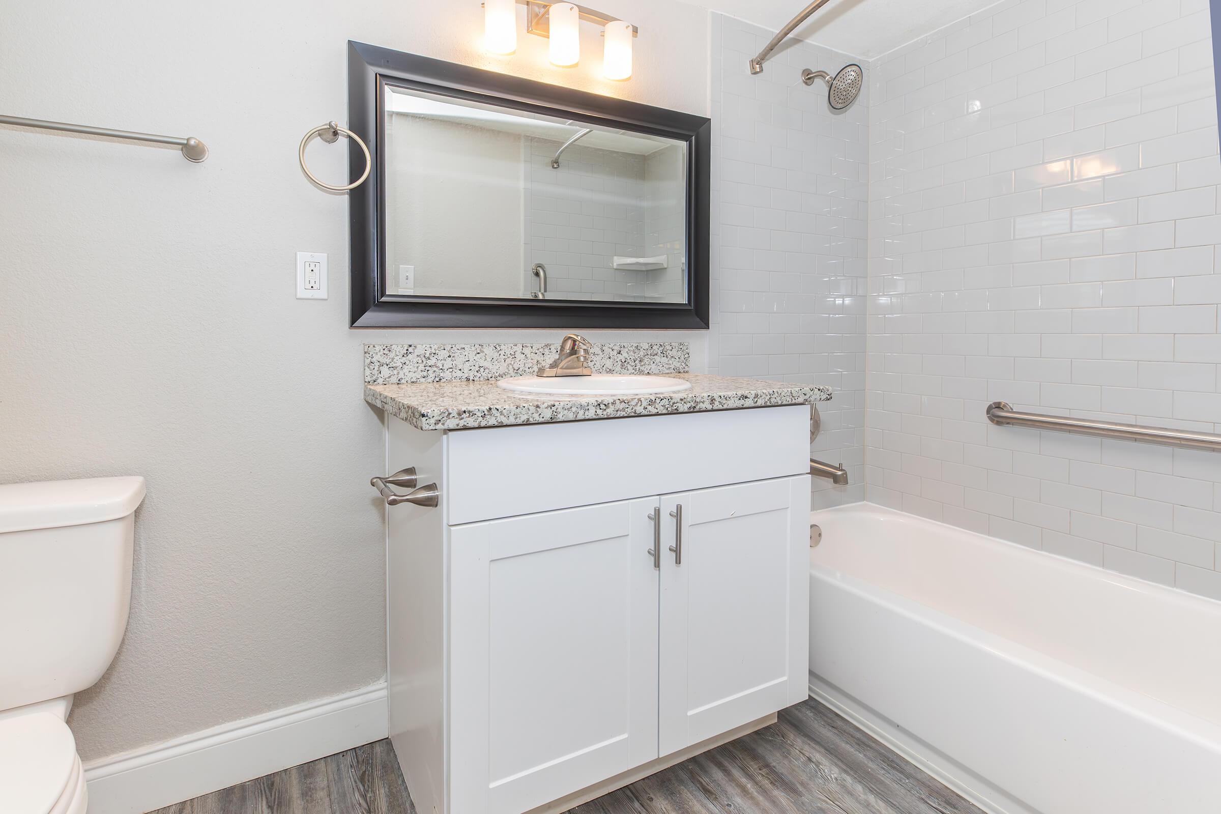 A modern bathroom featuring a white vanity with a granite countertop, a large mirror with overhead lighting, a tub with a shower, and gray tiled walls. The floor has a wood-like appearance, and there are towel bars and grab bars for convenience. The design is clean and contemporary.