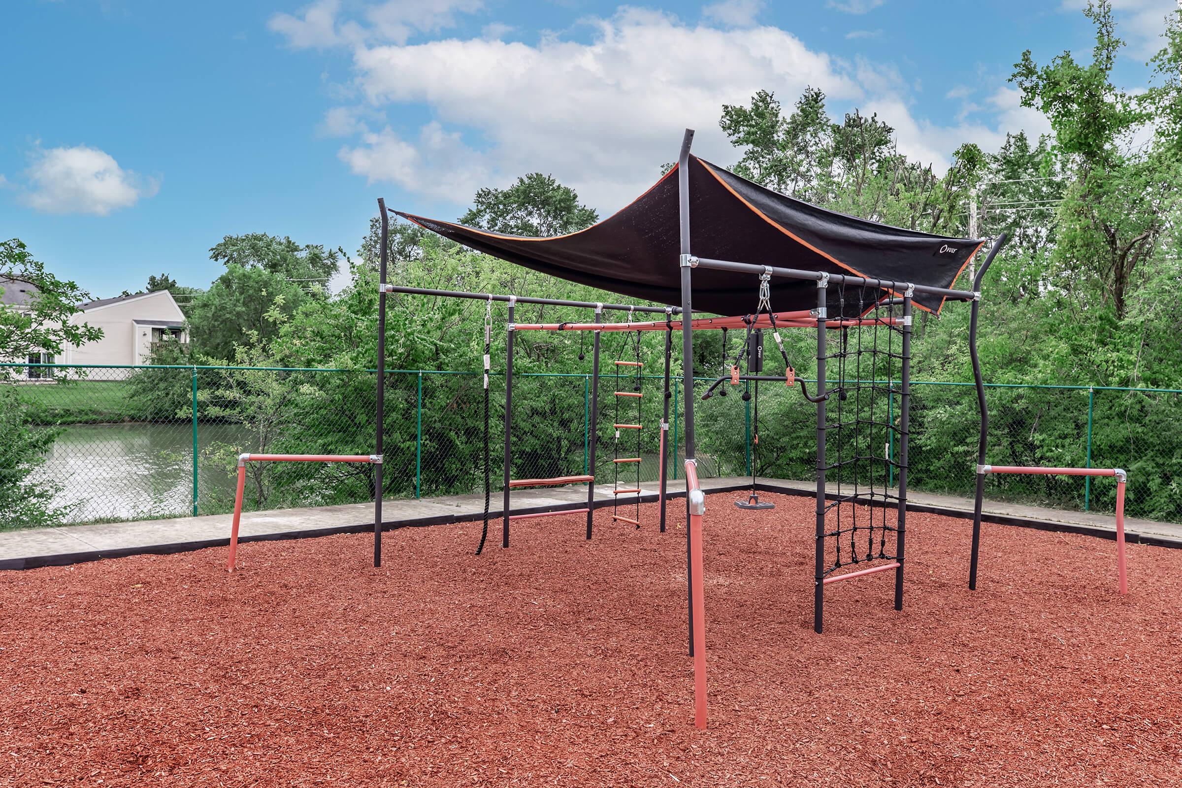 A playground structure with a black canopy, featuring climbing bars, swings, and a slide, situated on a bed of red mulch. Surrounding greenery and a fence are visible, with a cloudy sky in the background, creating a recreational outdoor space.