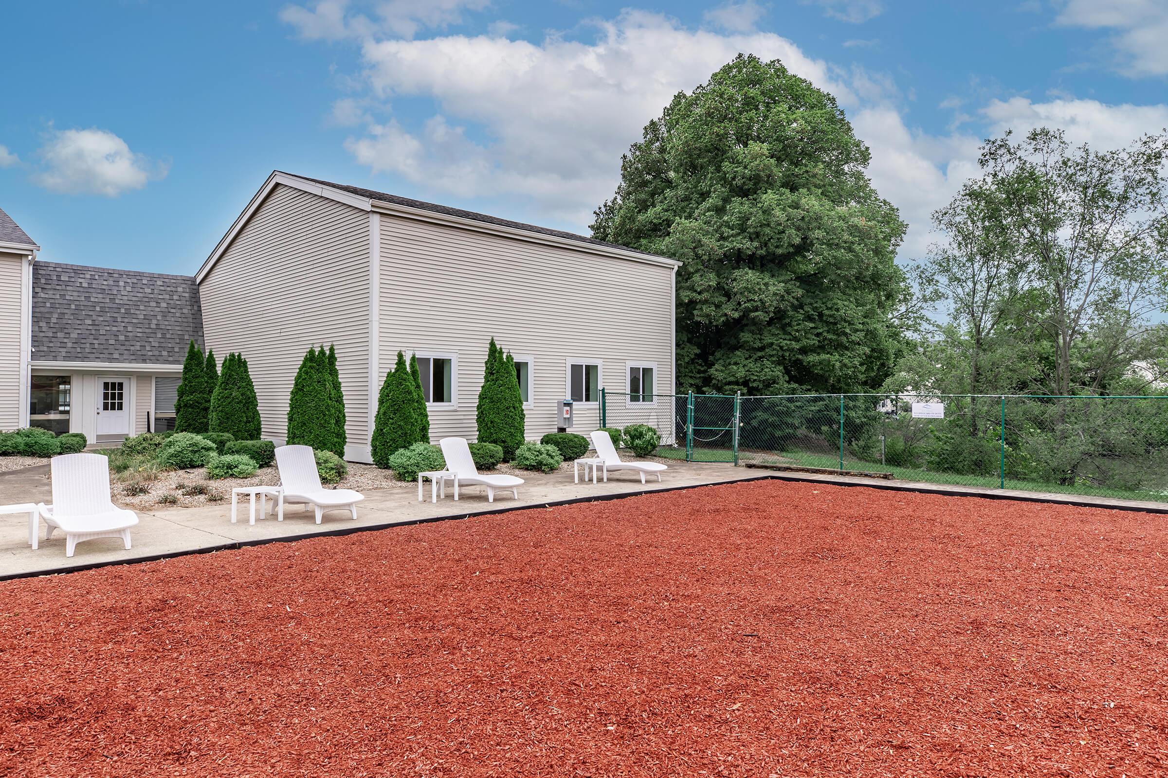 A landscaped area featuring red mulch, white lounge chairs, and a fenced space adjacent to a building. The background includes green bushes and trees under a partly cloudy sky.