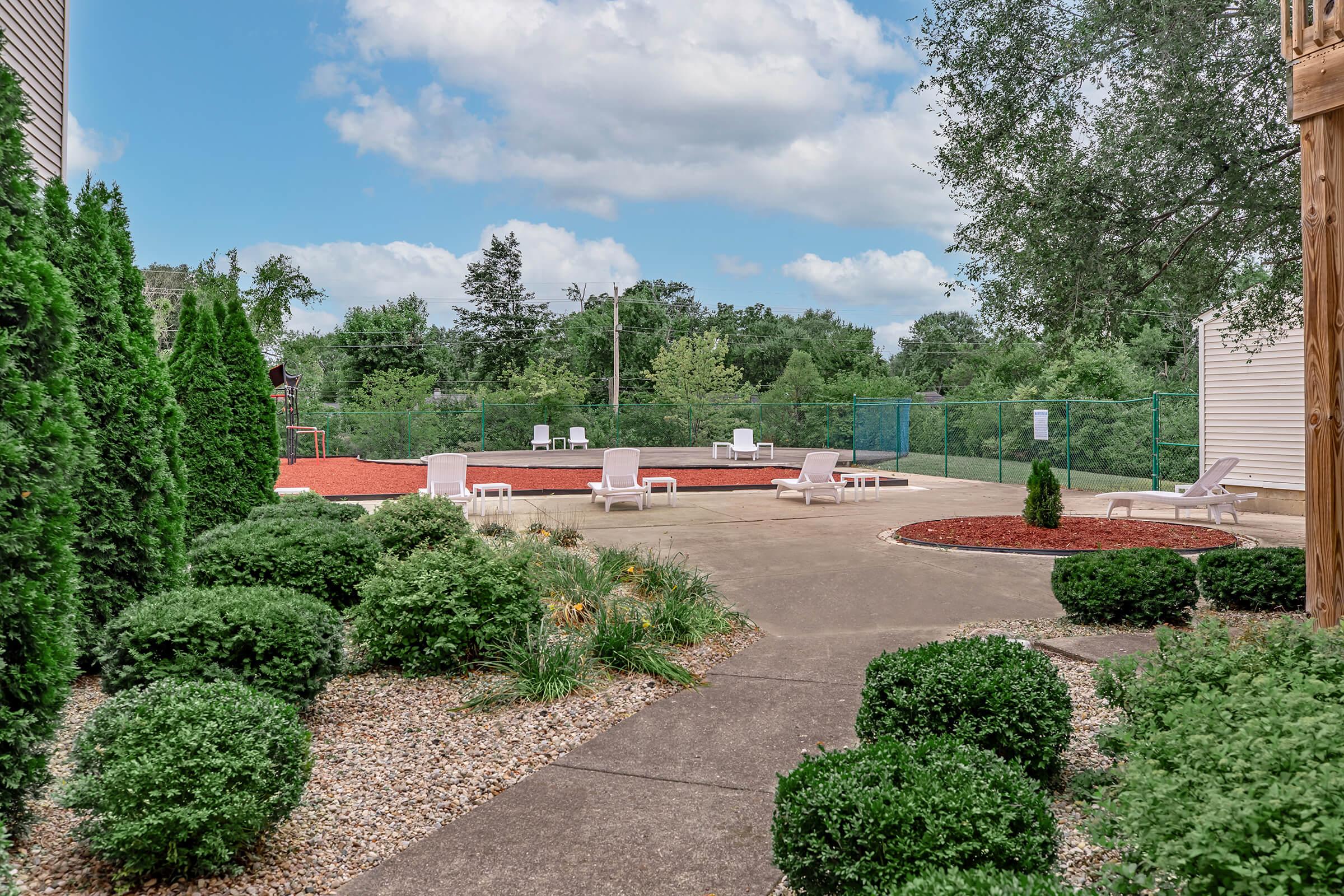 A landscaped outdoor area featuring a swimming pool surrounded by lounge chairs, with a red mulch play area in the background. Lush greenery and neatly trimmed bushes frame the scene, set against a bright blue sky with scattered clouds.