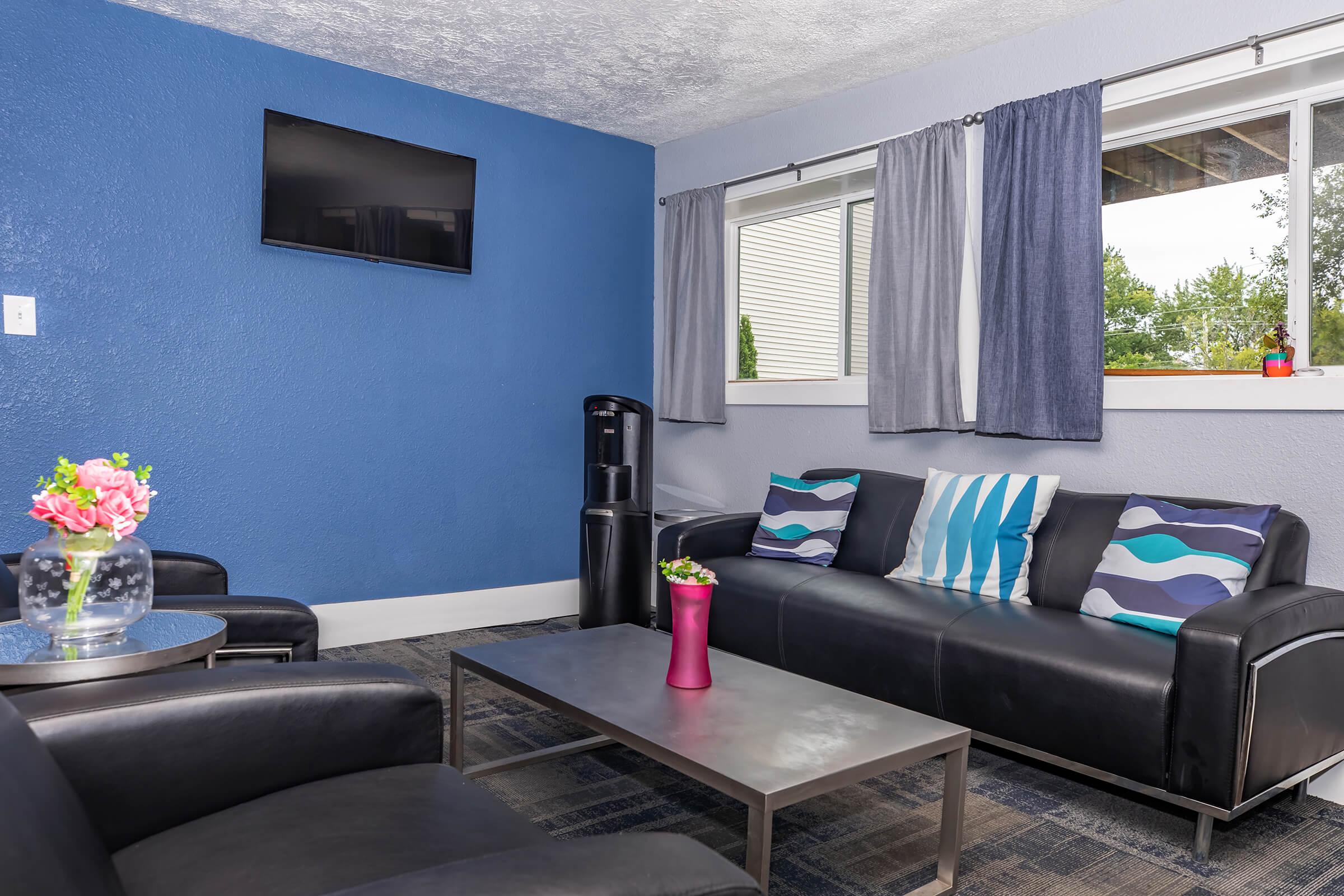 A cozy living area featuring a modern black leather sofa with decorative cushions, a coffee table, a small vase of flowers, and a water cooler. The walls are painted a deep blue, contrasted by light gray curtains hanging over a window that lets in natural light. A flat-screen TV is mounted on the wall.