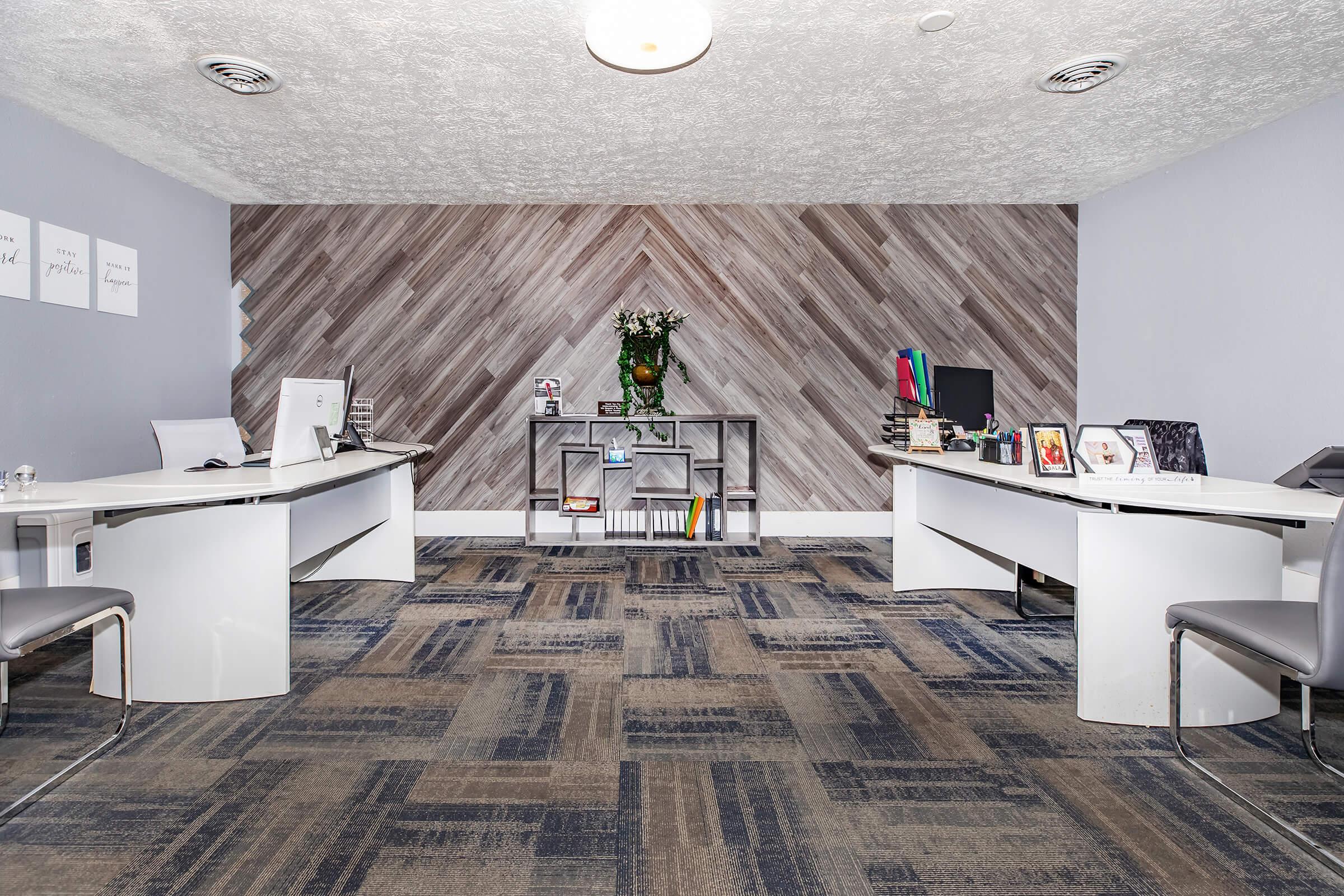 A modern office space featuring two white desks with computer monitors, stylish chairs, and a bookshelf. The wall has a wood-patterned design and a decorative plant in the center. The flooring is carpeted with a textured pattern, creating a professional and inviting atmosphere.