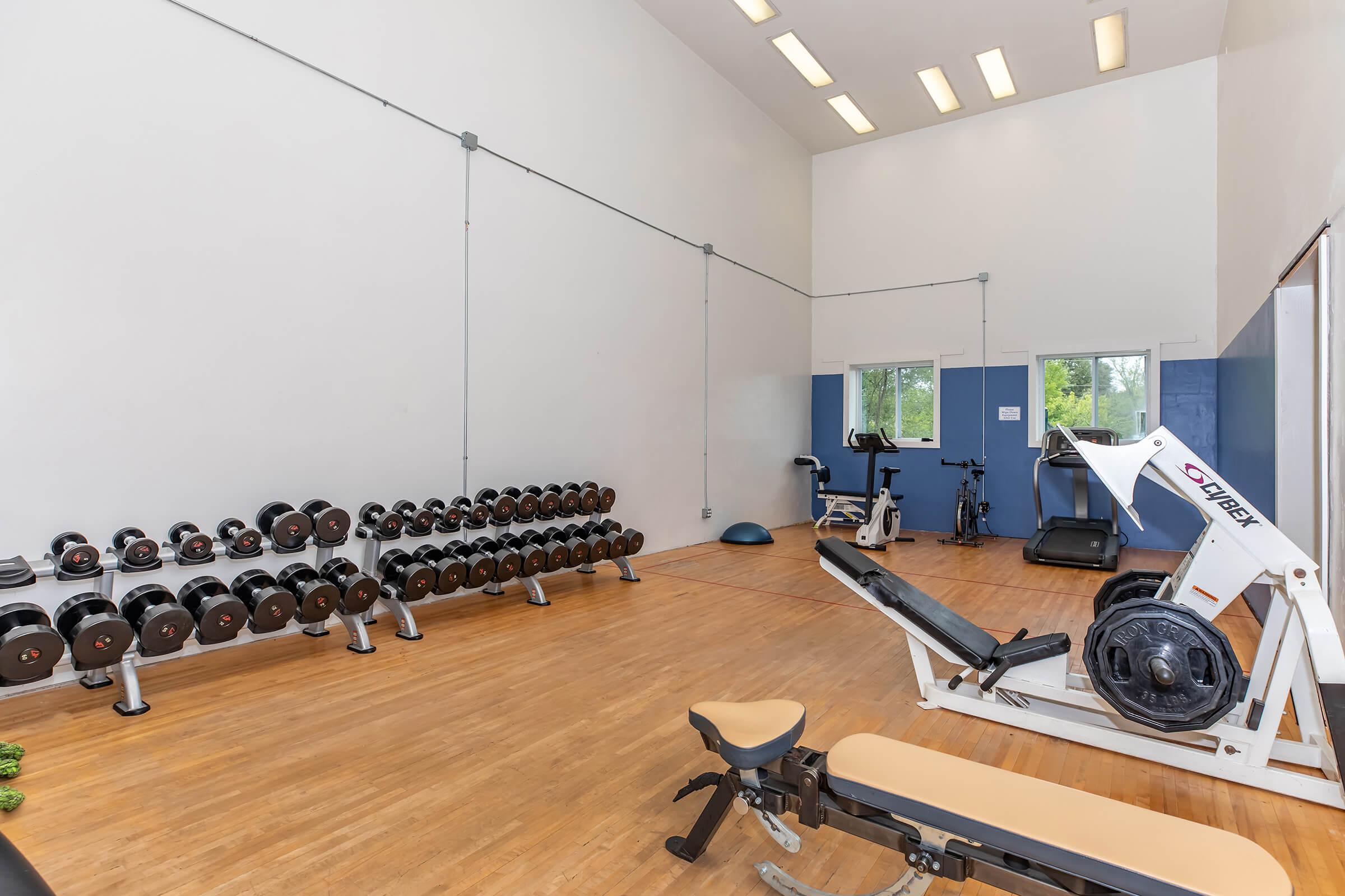 A bright and spacious fitness room featuring a rack of dumbbells, a weight bench, and a multi-use gym machine. There are also exercise bikes against the wall, and large windows allowing natural light to fill the space, enhancing the workout atmosphere. The floor is hardwood, adding to the clean aesthetic.