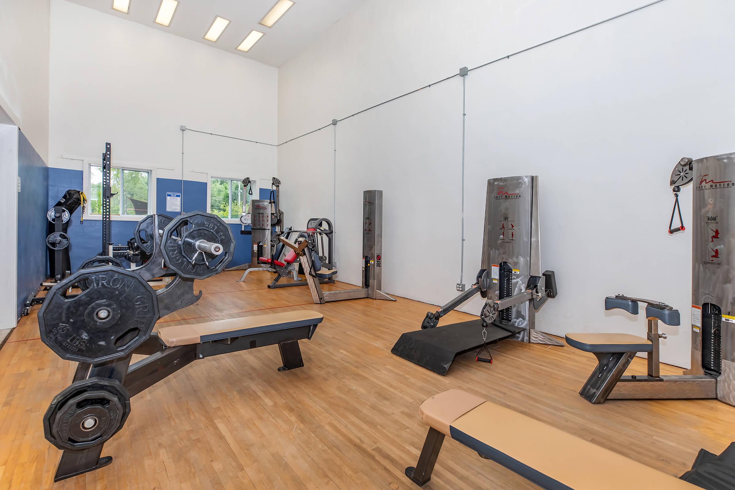 A bright and spacious gym interior featuring various weightlifting machines and benches. The wooden floor, white walls, and large windows create a welcoming atmosphere for exercise. Equipment includes a leg press machine, benches, and free weights, suitable for strength training.
