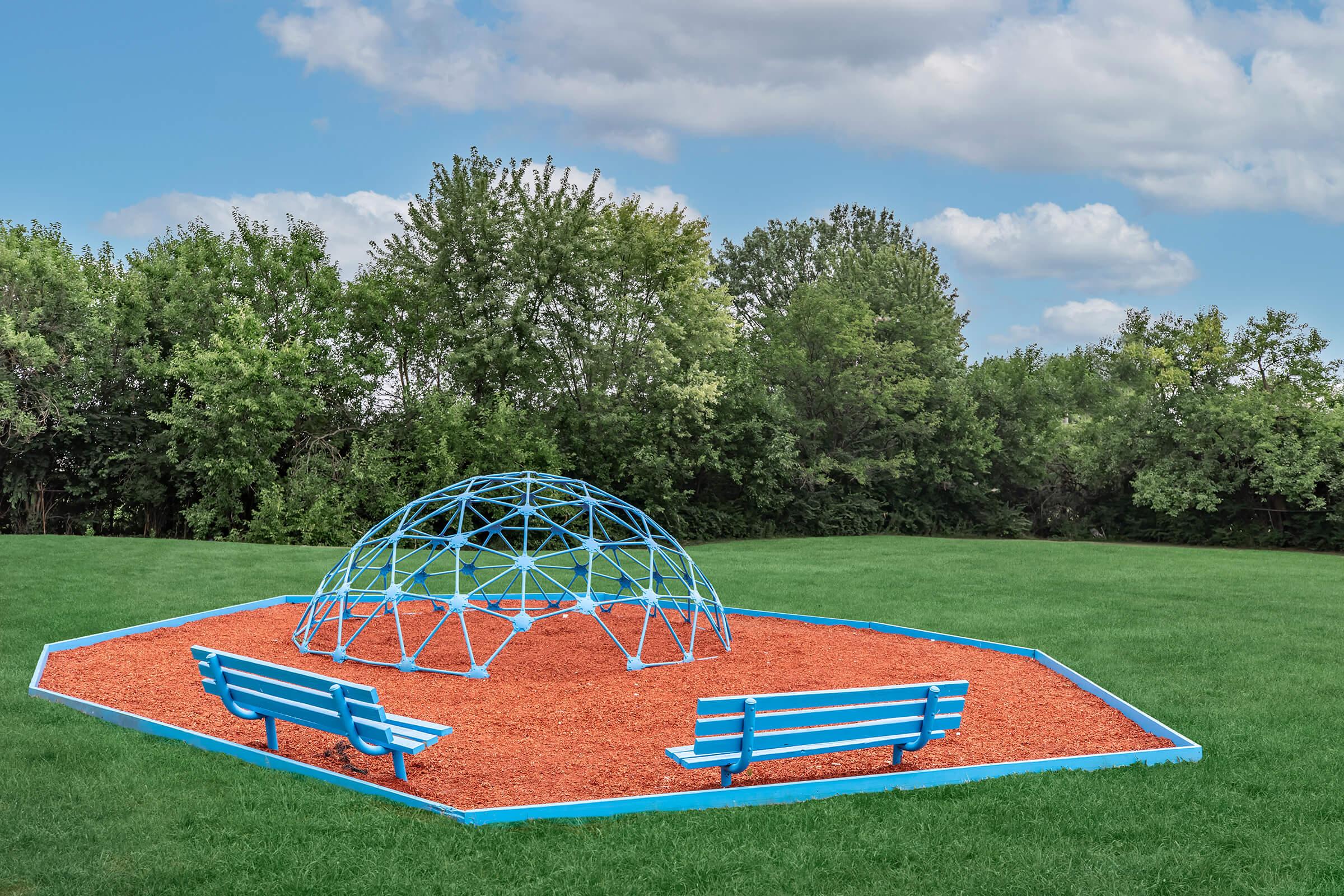 A blue geometric climbing structure surrounded by red mulch in a grassy area, with two blue benches nearby. The background features trees under a partly cloudy sky.