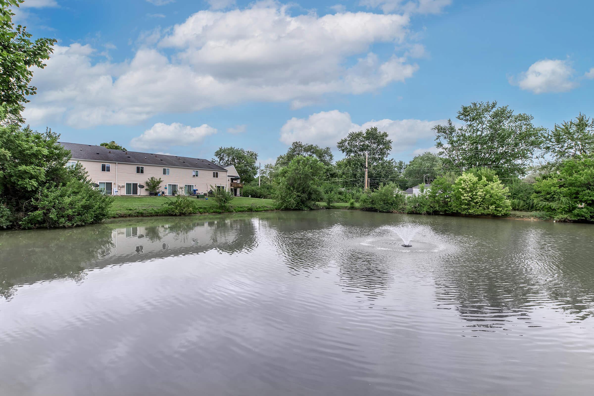 A serene pond surrounded by lush greenery and residential buildings. A fountain in the center creates ripples on the water's surface, reflecting the cloudy sky above. The landscape showcases a tranquil natural environment ideal for relaxation and leisure.