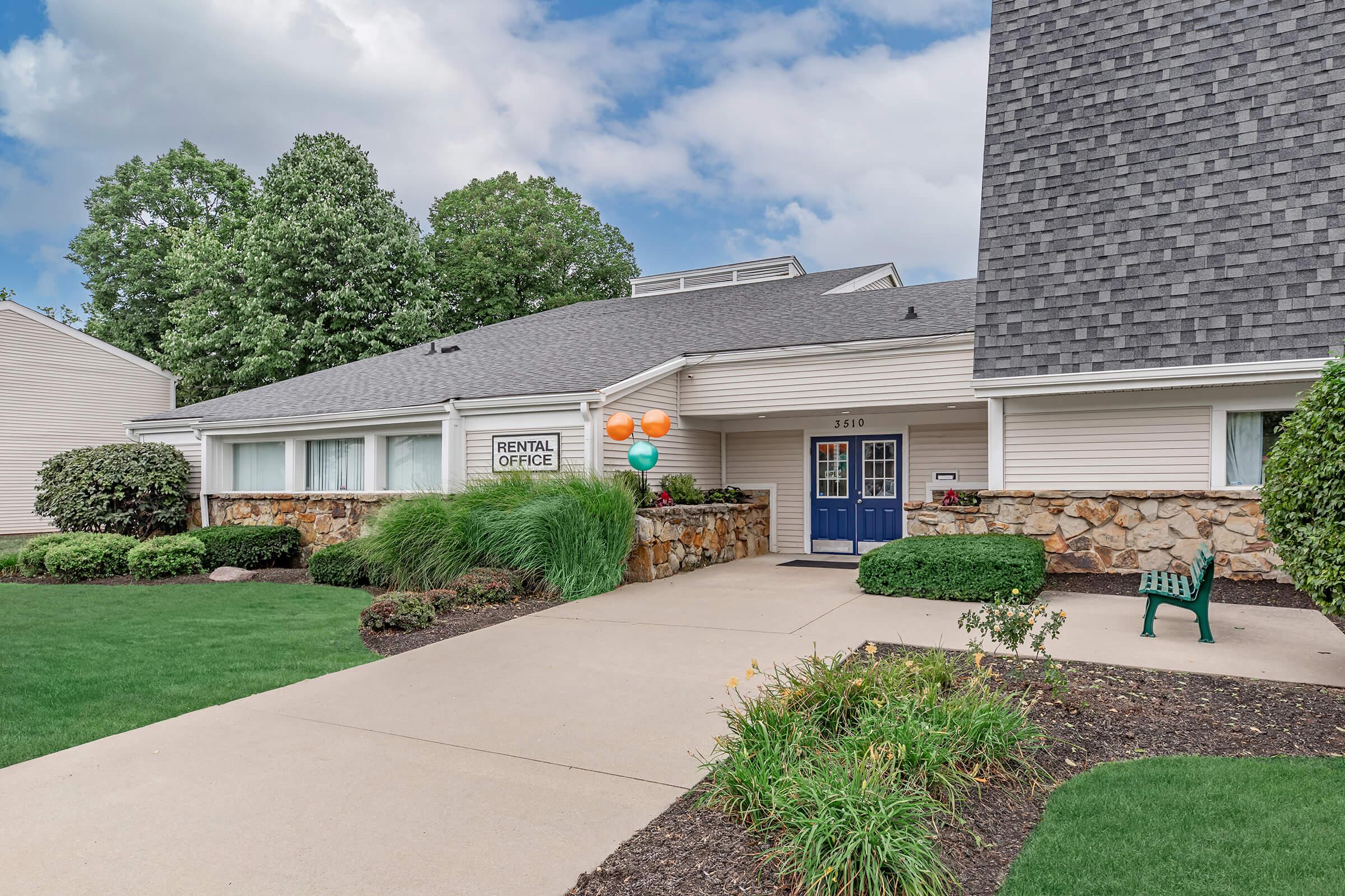 A rental office with a stone facade and blue door, surrounded by manicured grass and shrubs. Colorful balloons are displayed near the entrance. The pathway leading to the office is paved, and a green bench can be seen in the foreground, with trees in the background under a cloudy sky.