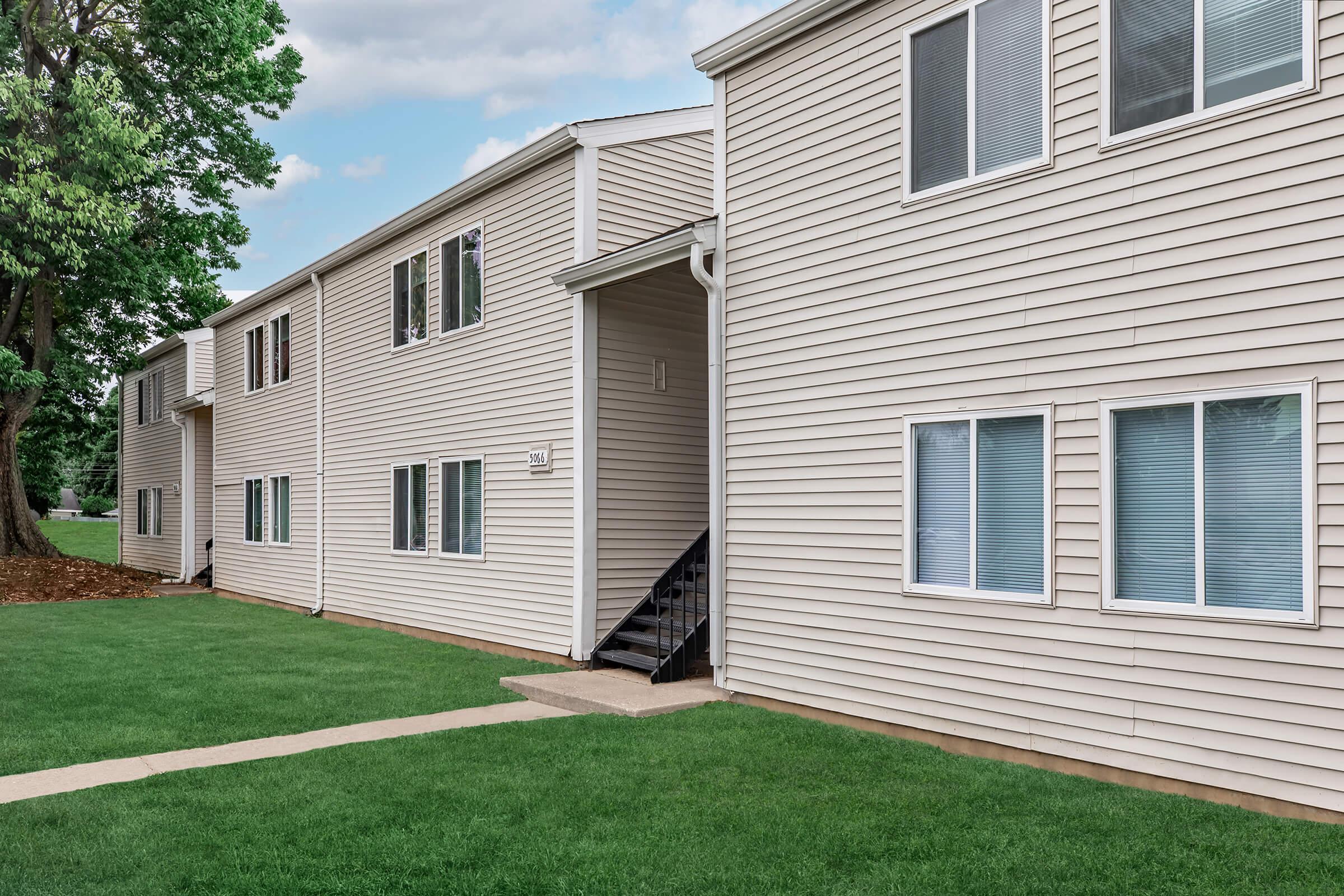 A two-story apartment building with beige vinyl siding, featuring a central staircase leading to the second floor. The grassy area in front is well-maintained, and several windows are visible on the façade. Trees are in the background, adding greenery to the scene.