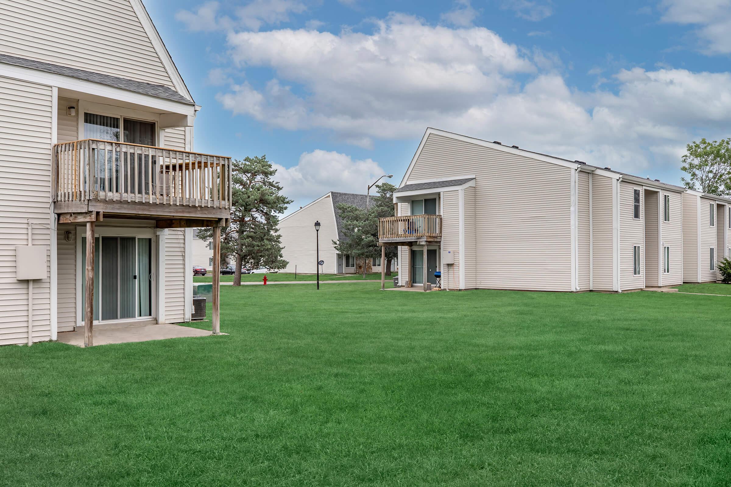 Two residential buildings with balconies on the left and right, situated on a well-maintained green lawn. In the background, additional buildings and trees are visible under a partly cloudy sky. The scene conveys a peaceful suburban environment.
