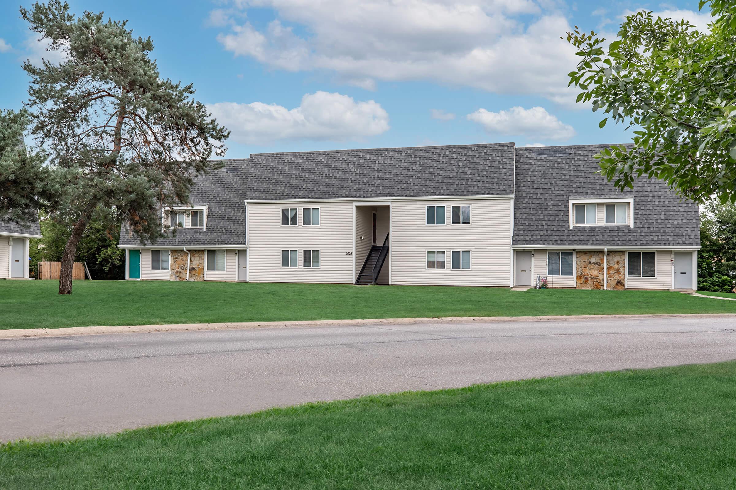 A view of a two-story residential building with a gray shingle roof and multiple windows. The building features a light-colored exterior and is set in a landscaped area with green grass. There are trees and a cloudy sky in the background. A pathway leads up to the entrance, which is visible in the center.