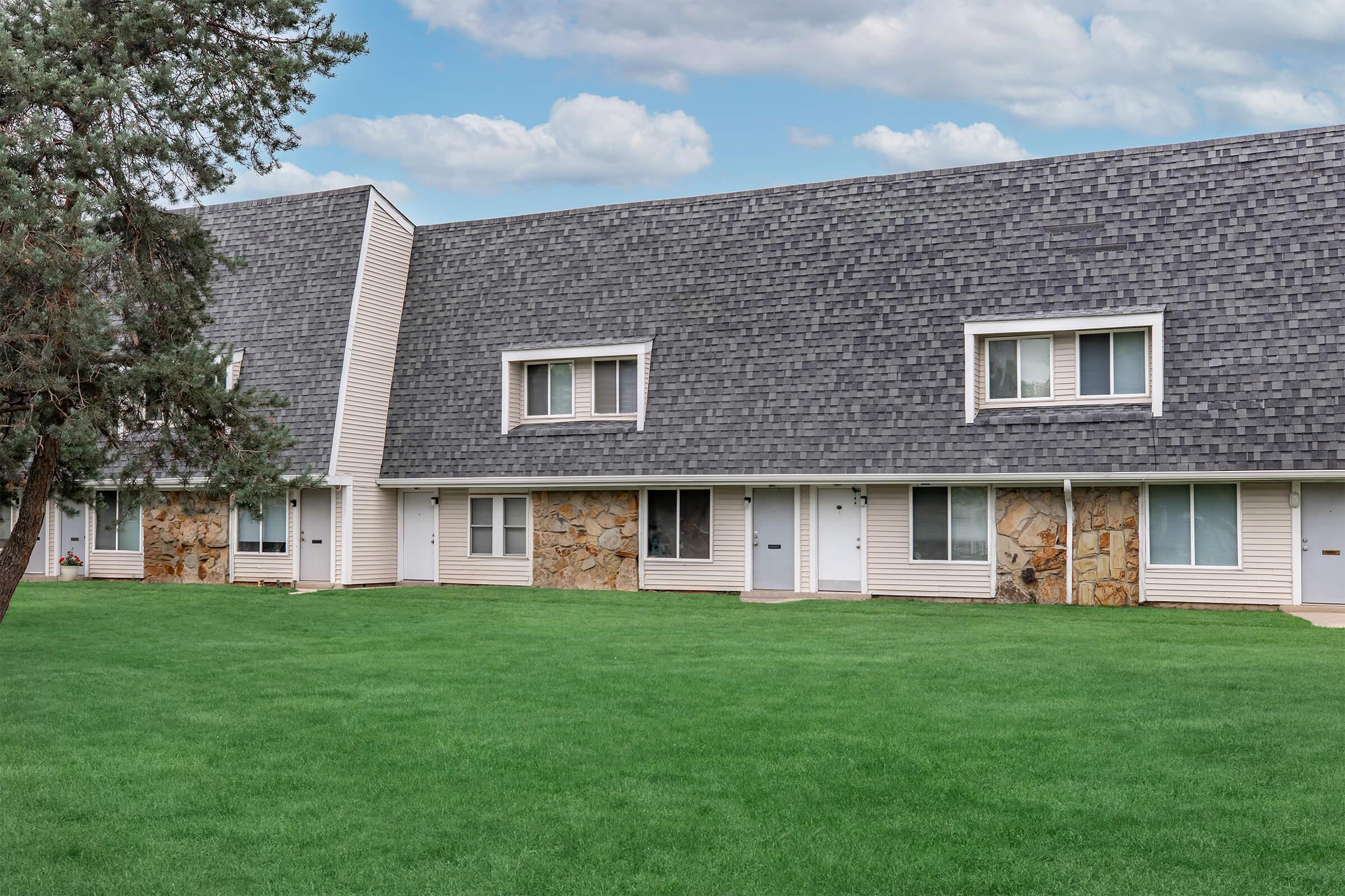 A row of multi-unit residential buildings with a sloped roof, featuring stone accents at the base. The green lawn in front is well-maintained, and the sky above is partly cloudy. Each unit has a porch area and windows, creating a pleasant suburban atmosphere.