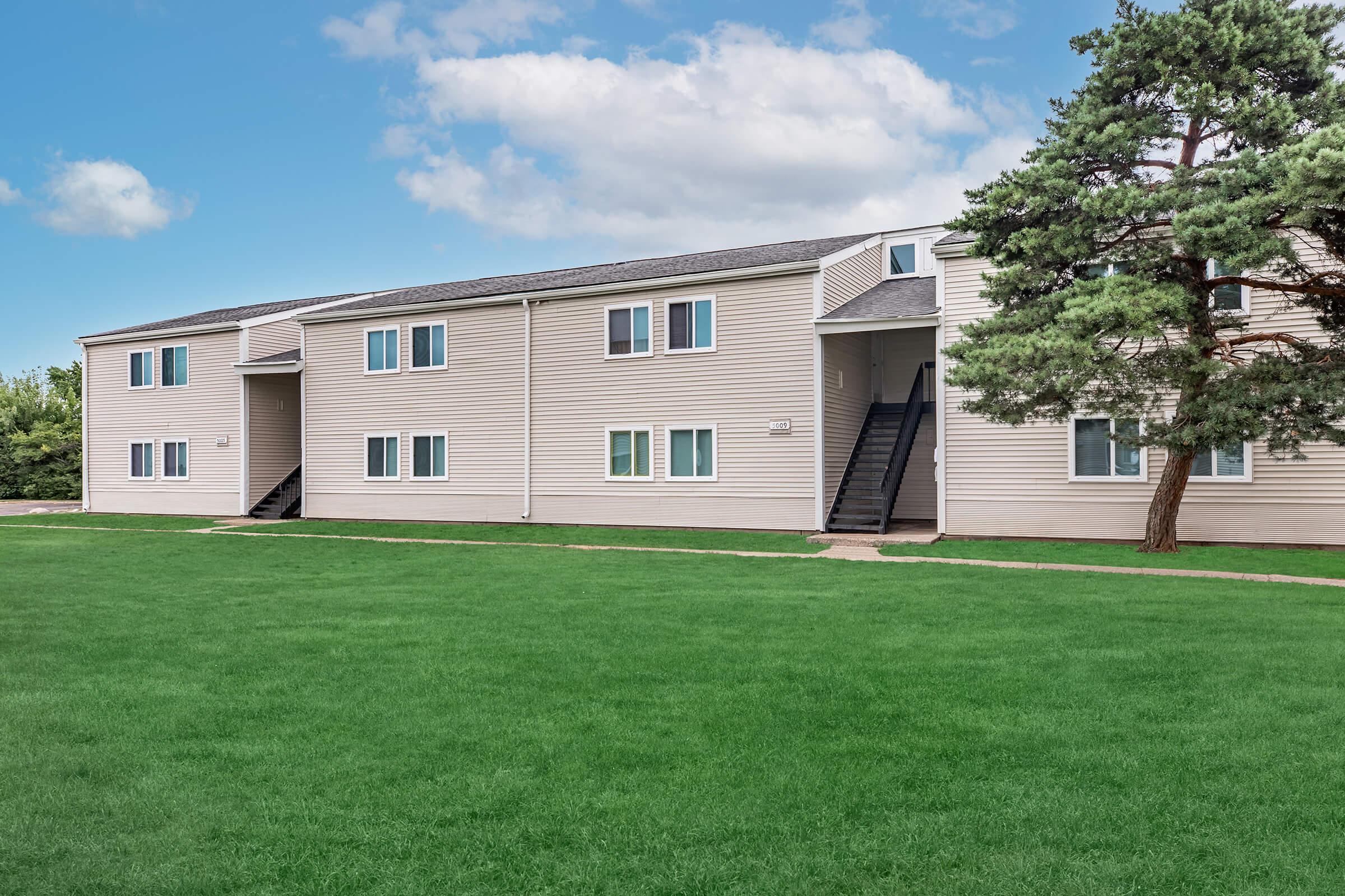 A multi-unit residential building with beige siding and multiple windows, set on a green lawn. The building features a staircase on one side and is surrounded by trees under a partly cloudy sky.
