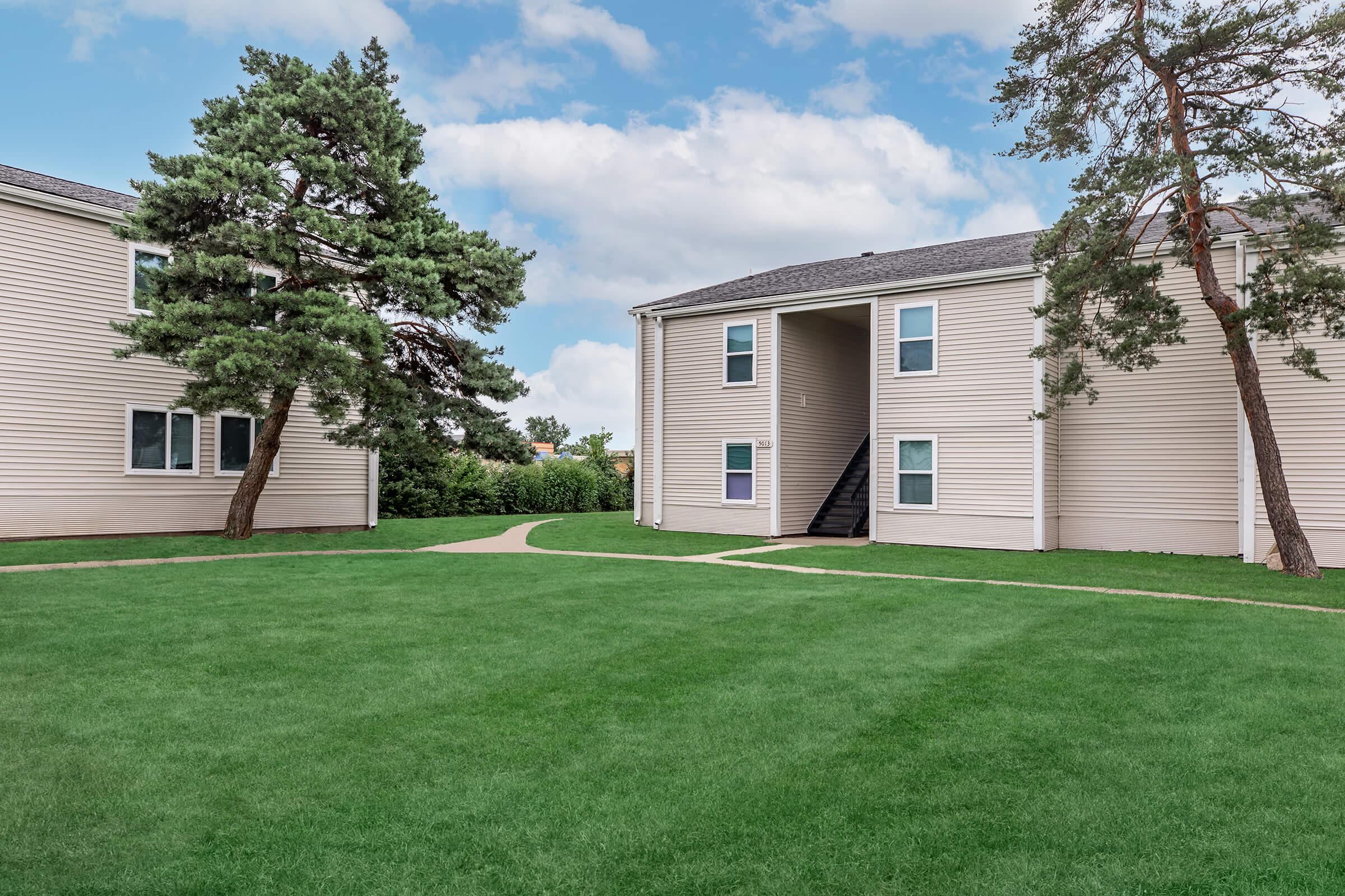 A view of a grassy courtyard between two residential buildings. The buildings are light-colored with balconies, and there are trees on either side providing some shade. The sky is partly cloudy, contributing to a serene atmosphere.